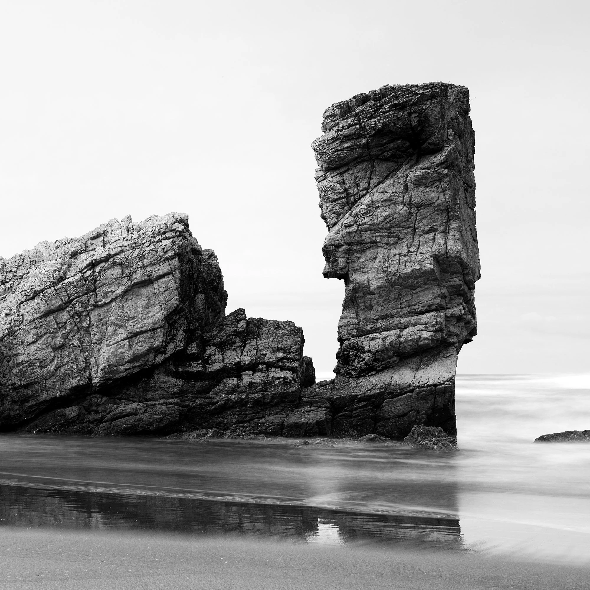© 2023 Gerald Berghammer. Black-and-white beach scene with wet sand, faint footprints, and a tall rock formation near the shore reflected in shallow water. Print detail 3