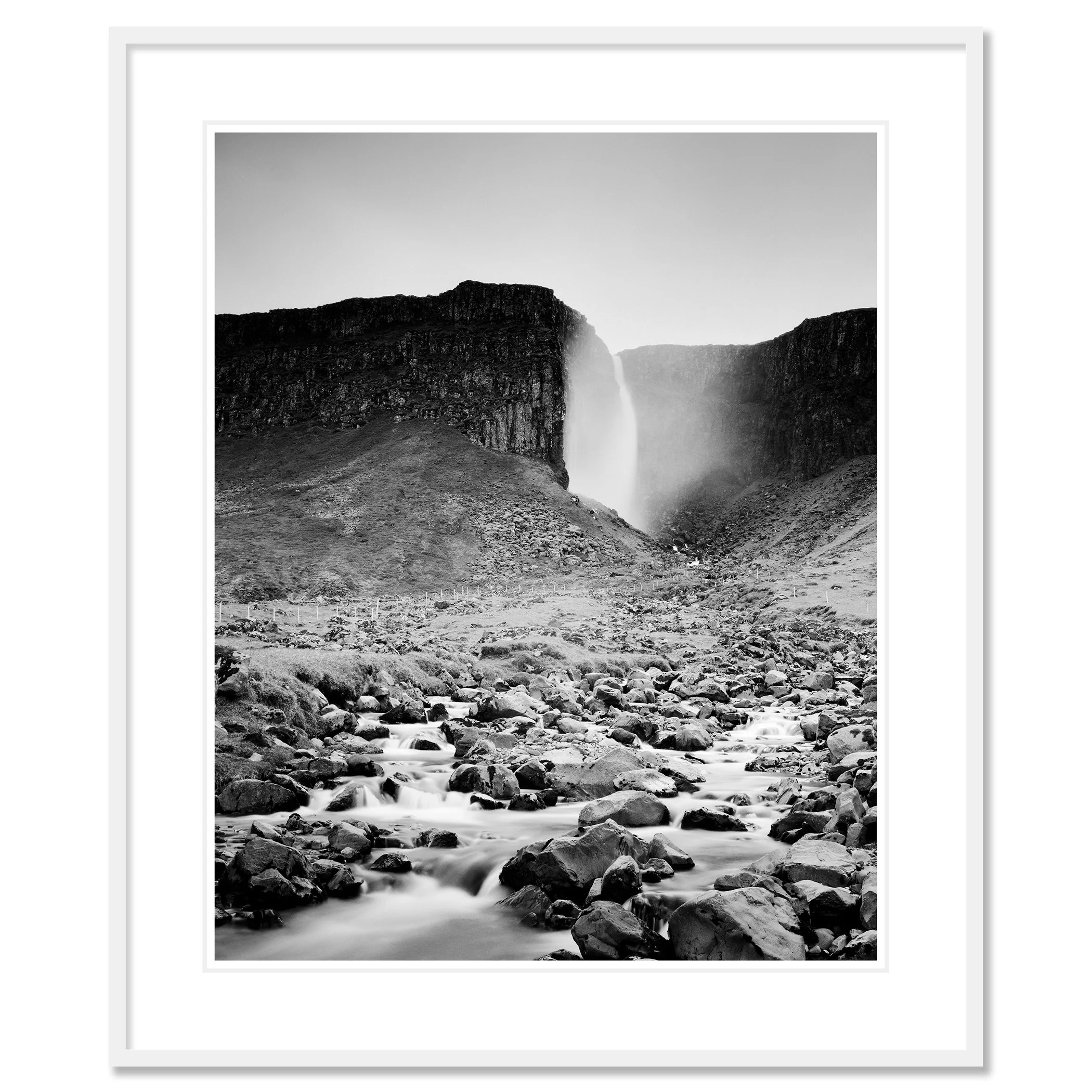 Gerald Berghammer - Black and white landscape photography. A waterfall cascading down a rocky cliff into a stream surrounded by rocks and rugged terrain. Classic framed white