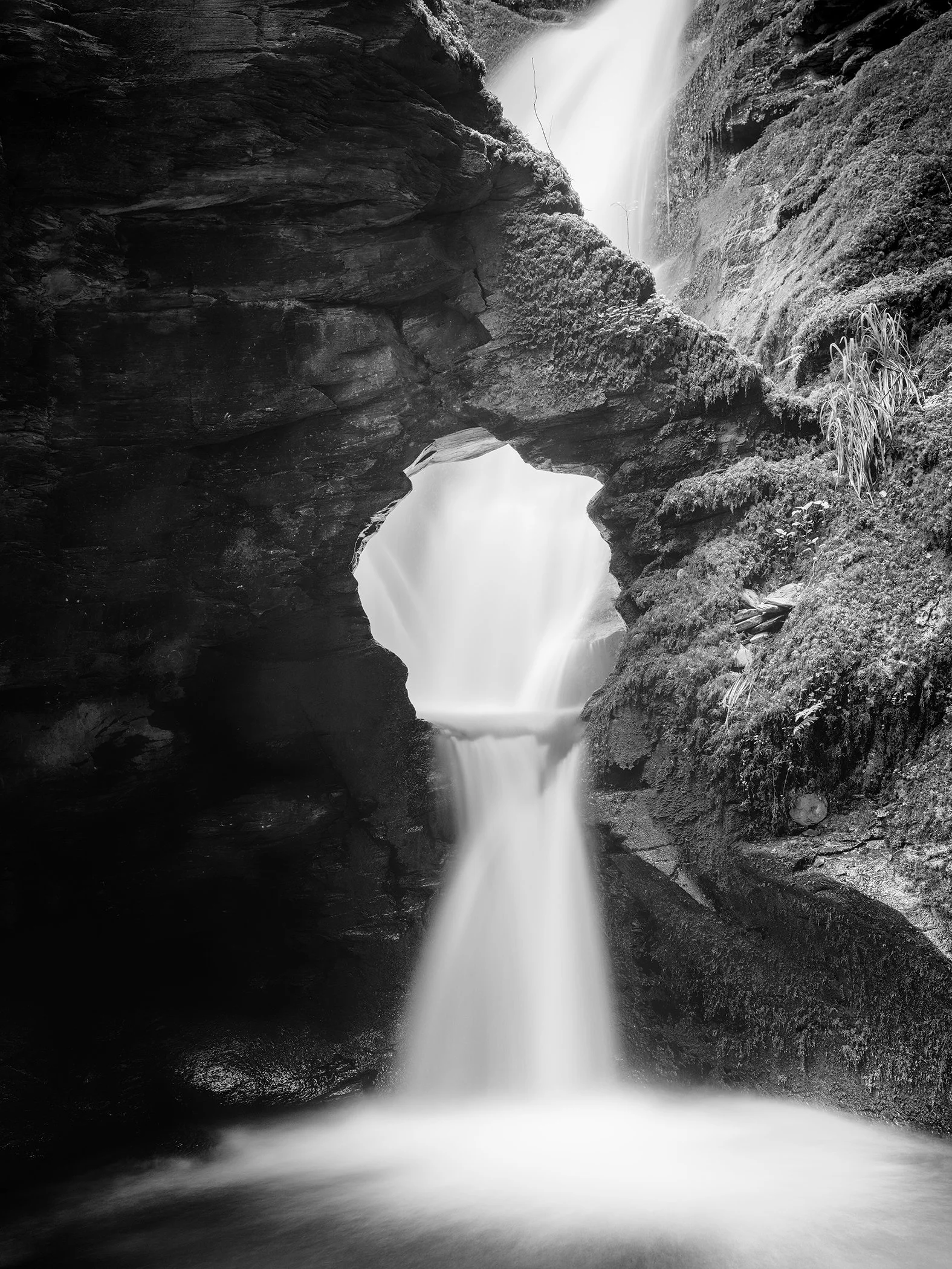 Black and white long exposure photograph of a waterfall flowing through a natural rock arch in a mossy gorge