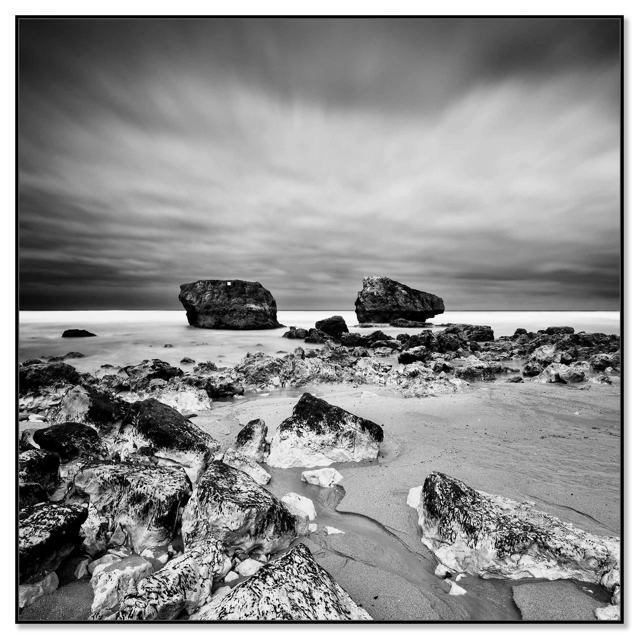 Black-and-white rocky beach with boulders offshore under dramatic, streaked clouds – framed ArtBox black