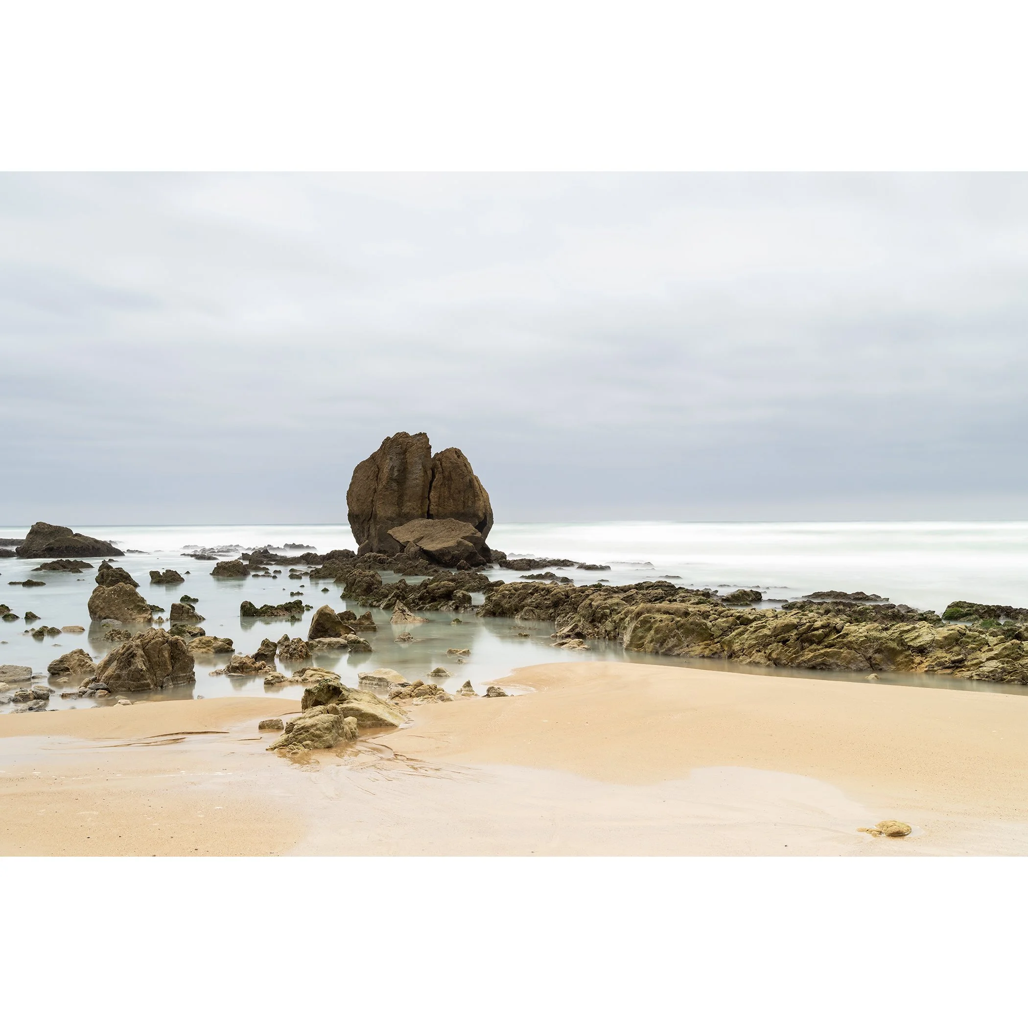 Gerald Berghammer - Color Seascape Photography. A beach with rocks and sand, with large rock formations rising from the water, under a cloudy sky.