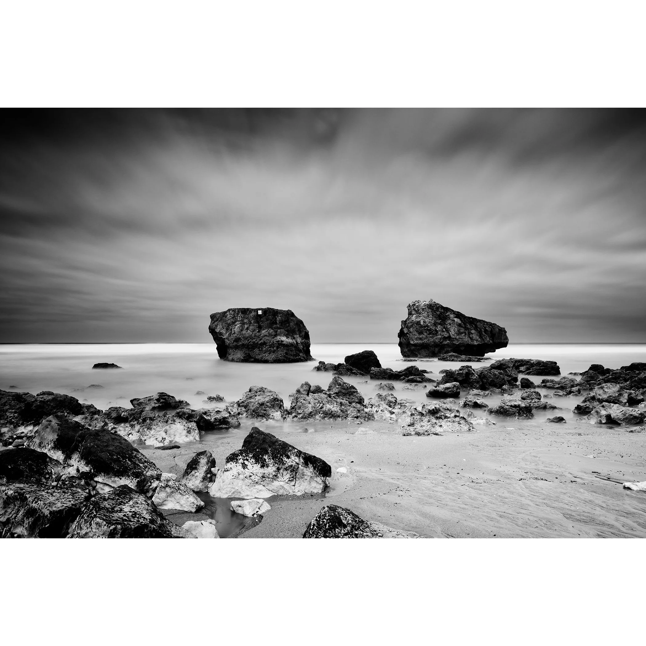 Gerald Berghammer - Black and white landscape photo. Rocky beach with two large rocks protruding from the water, cloudy sky, and smooth ocean surface.