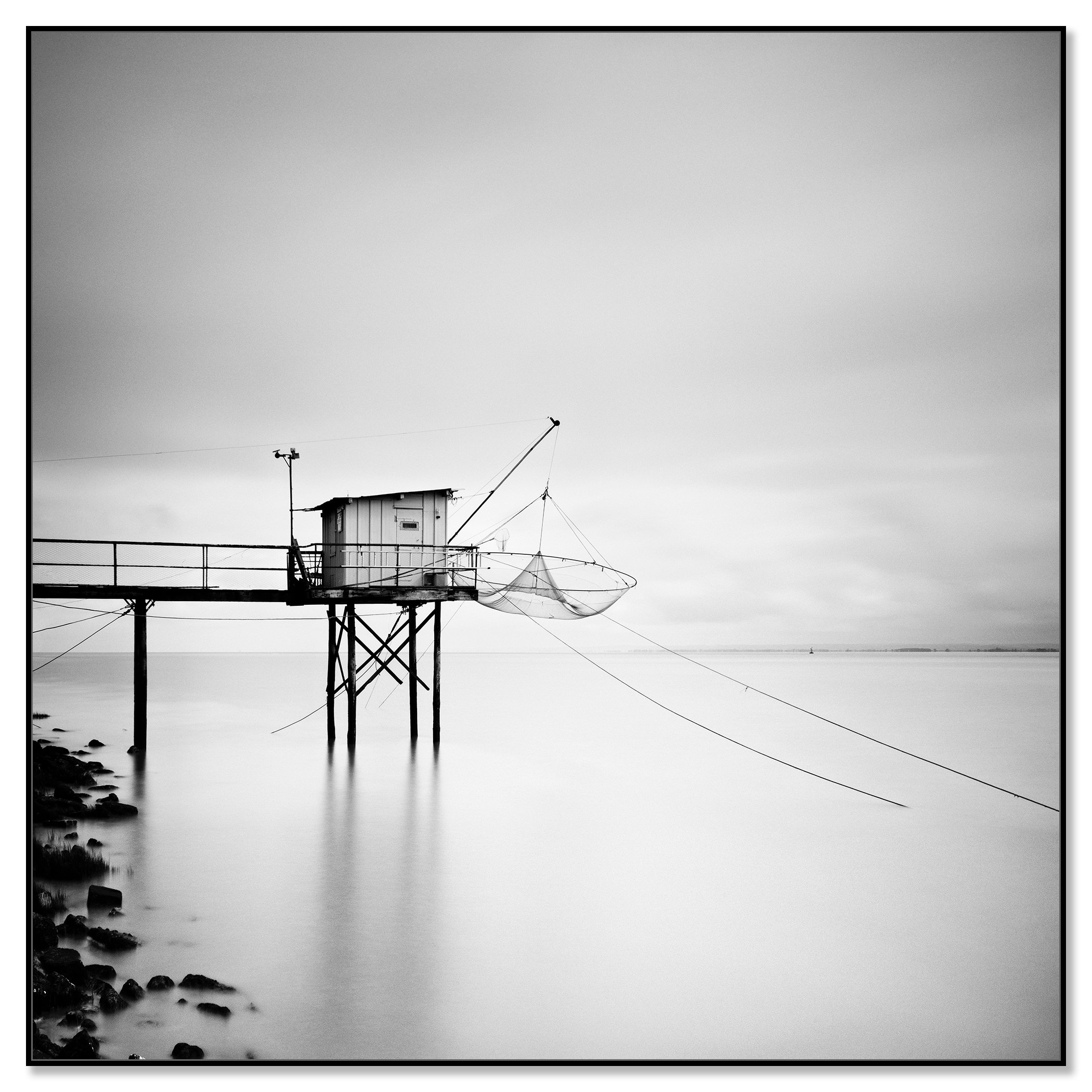 Black-and-white photo of a stilted fishing hut with a net over calm water beside a rocky shoreline – framed ArtBox black