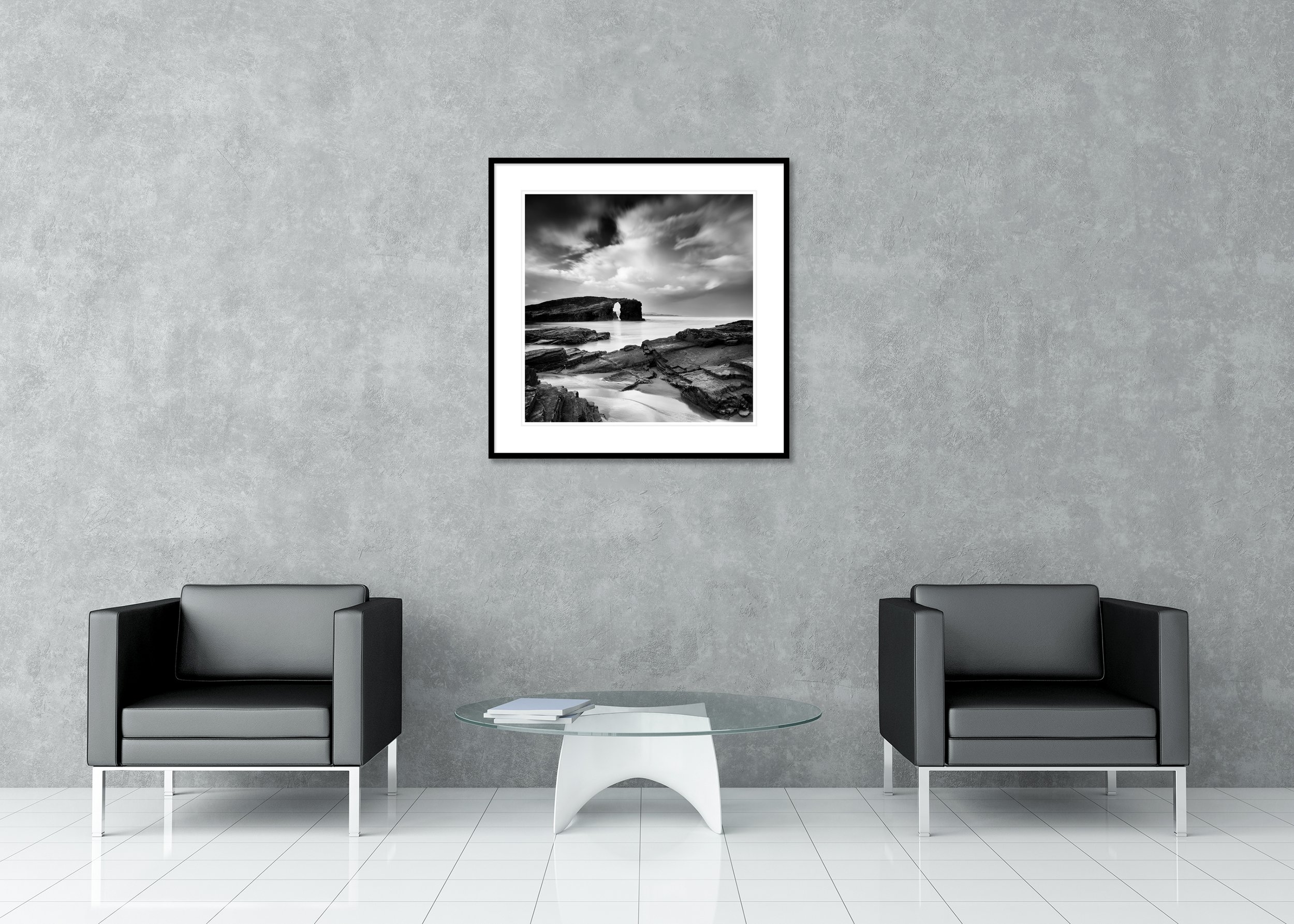 Black and white archival pigment print of a rocky beach with a natural sea arch, calm water and dark clouds, framed with a classic mount