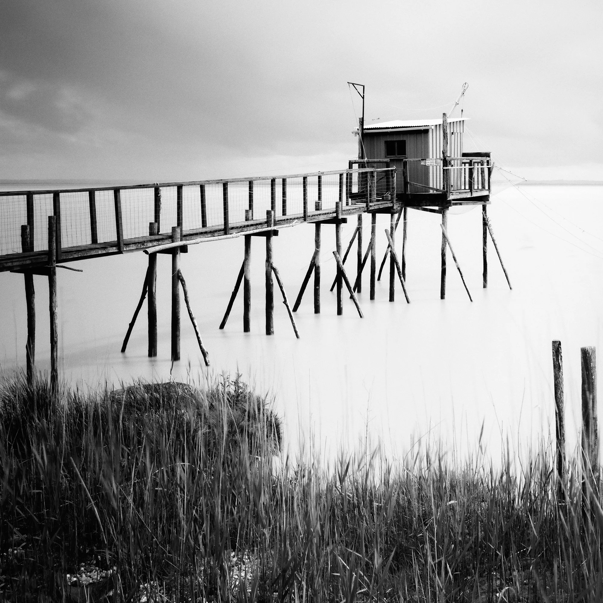 Gerald Berghammer - Black and white seascape photography. Small wooden hut on stilts connected by a long walkway over water with grass and a cloudy sky. Print detail 1