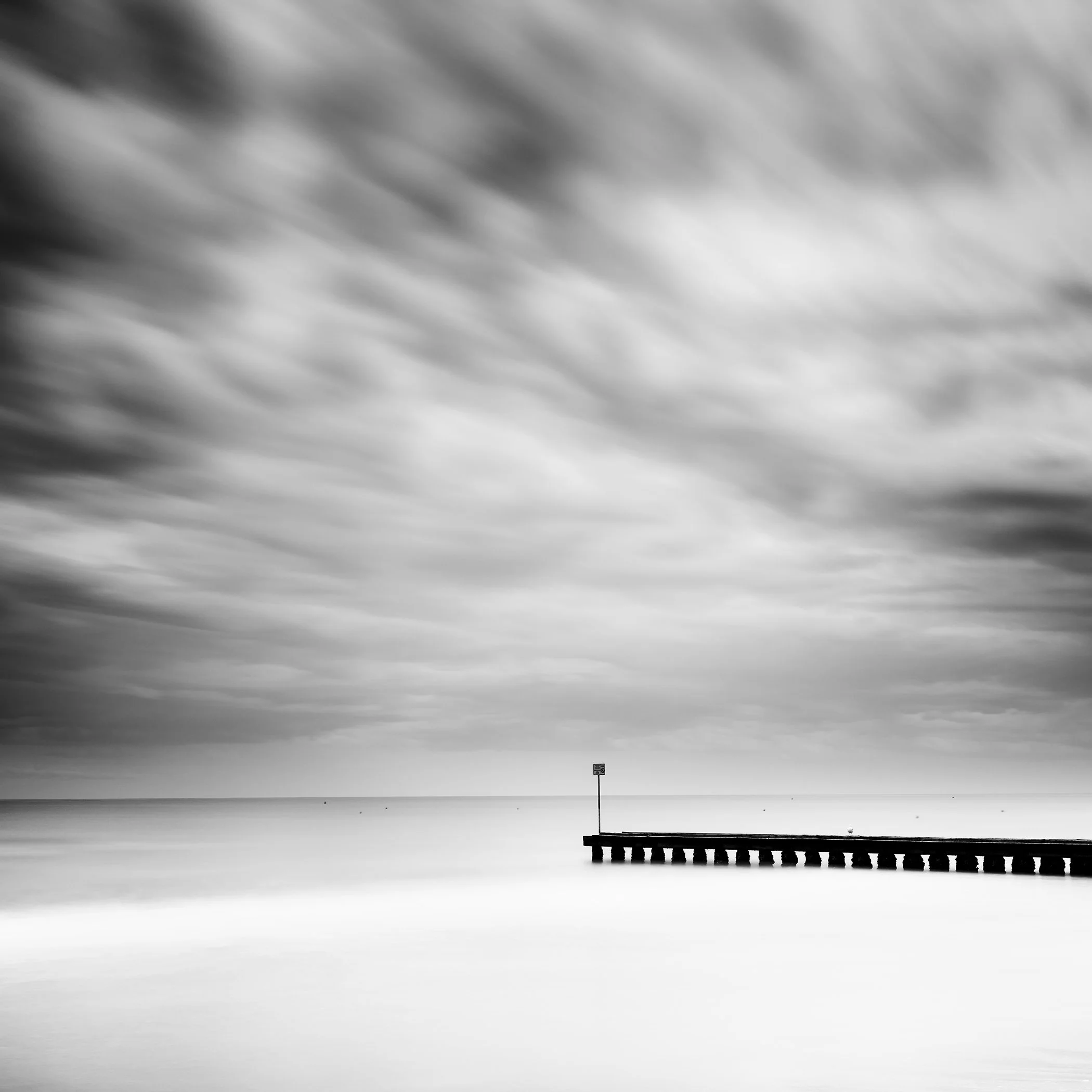 Wooden Pier in the Sea Study 1 – minimalist black-and-white long-exposure seascape with a wooden pier beneath dramatic clouds in Italy by Gerald Berghammer.