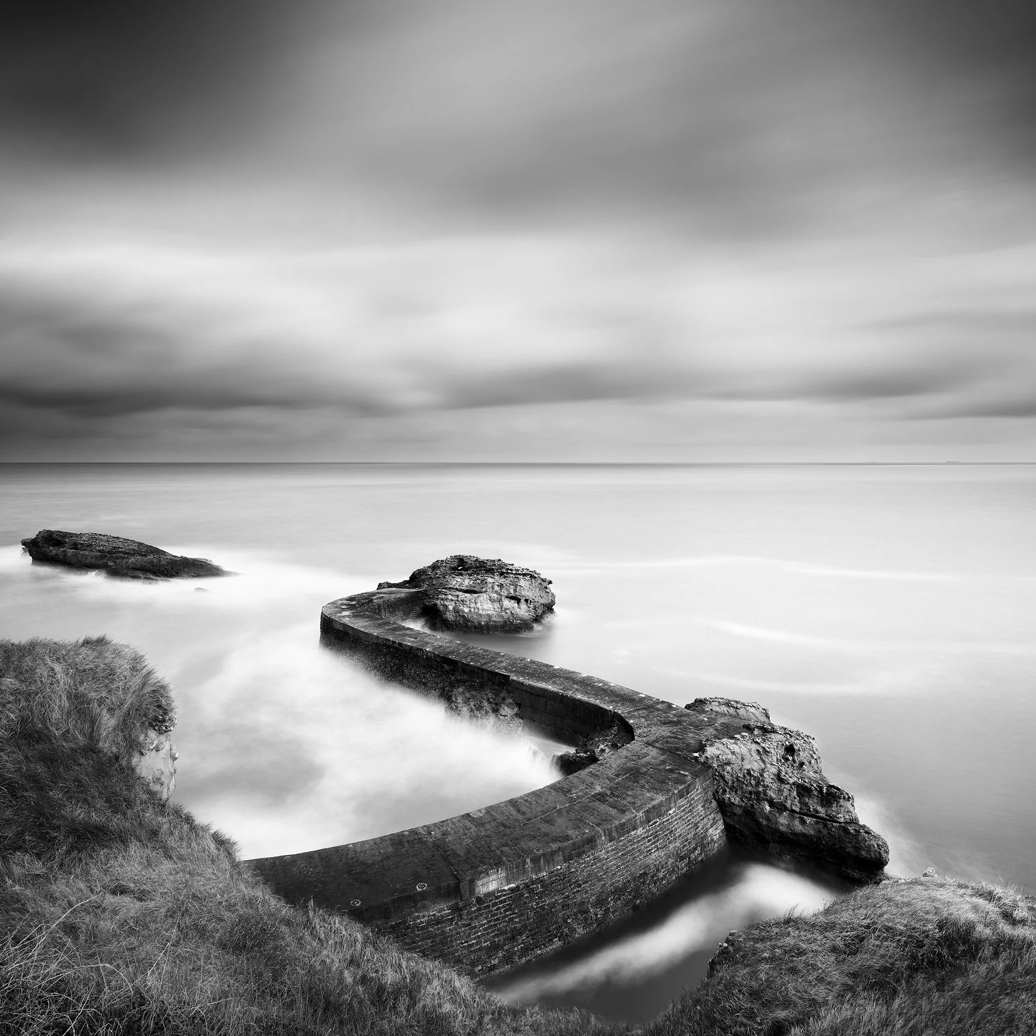 Breakwater Pier Study 1 by Gerald Berghammer, black-and-white long-exposure seascape of a curved stone breakwater on the French Atlantic coast.