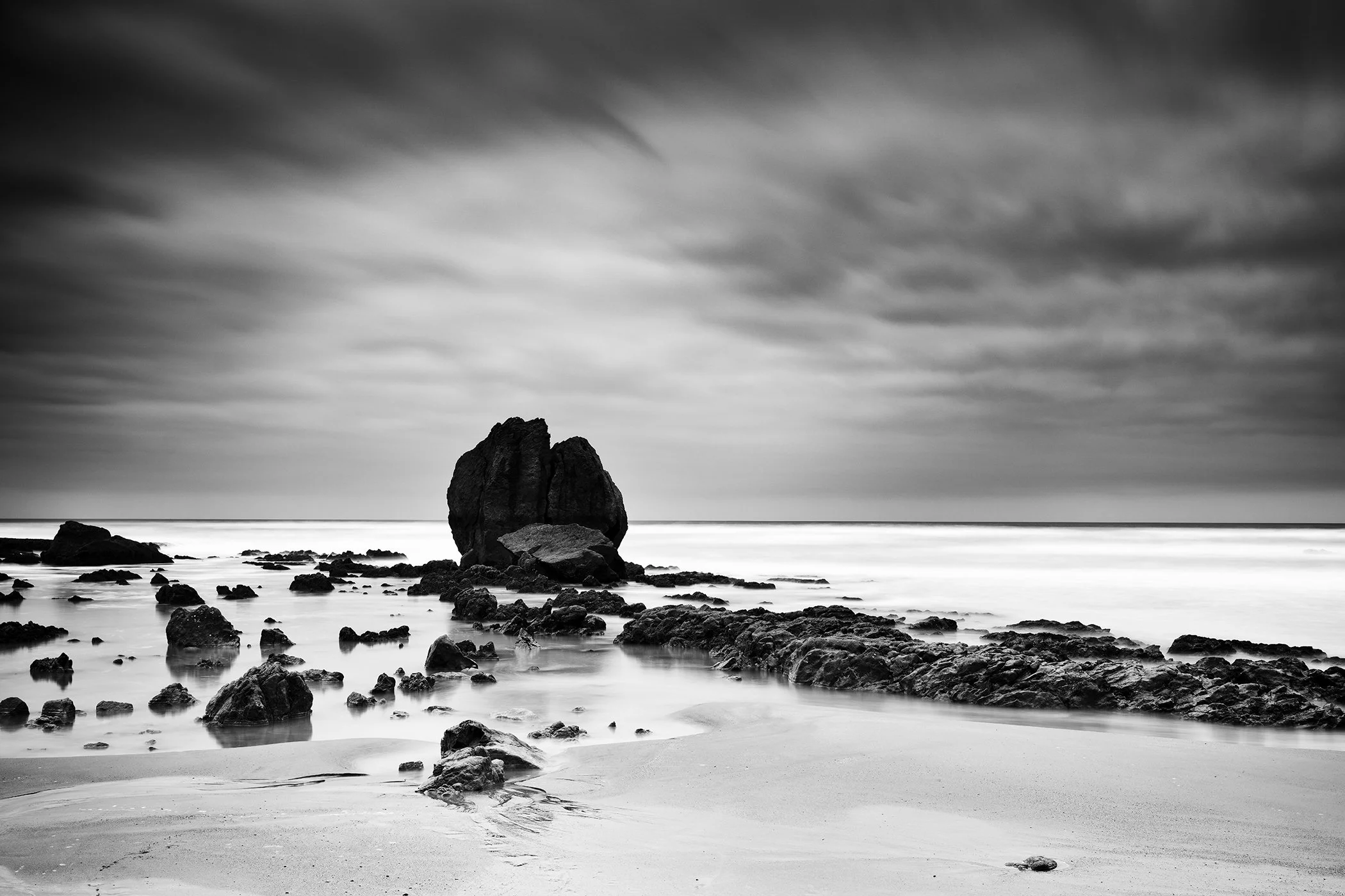 Long-exposure monochrome photo of a rocky beach with soft waves and an overcast sky, creating a moody seascape by Gerald Berghammer.