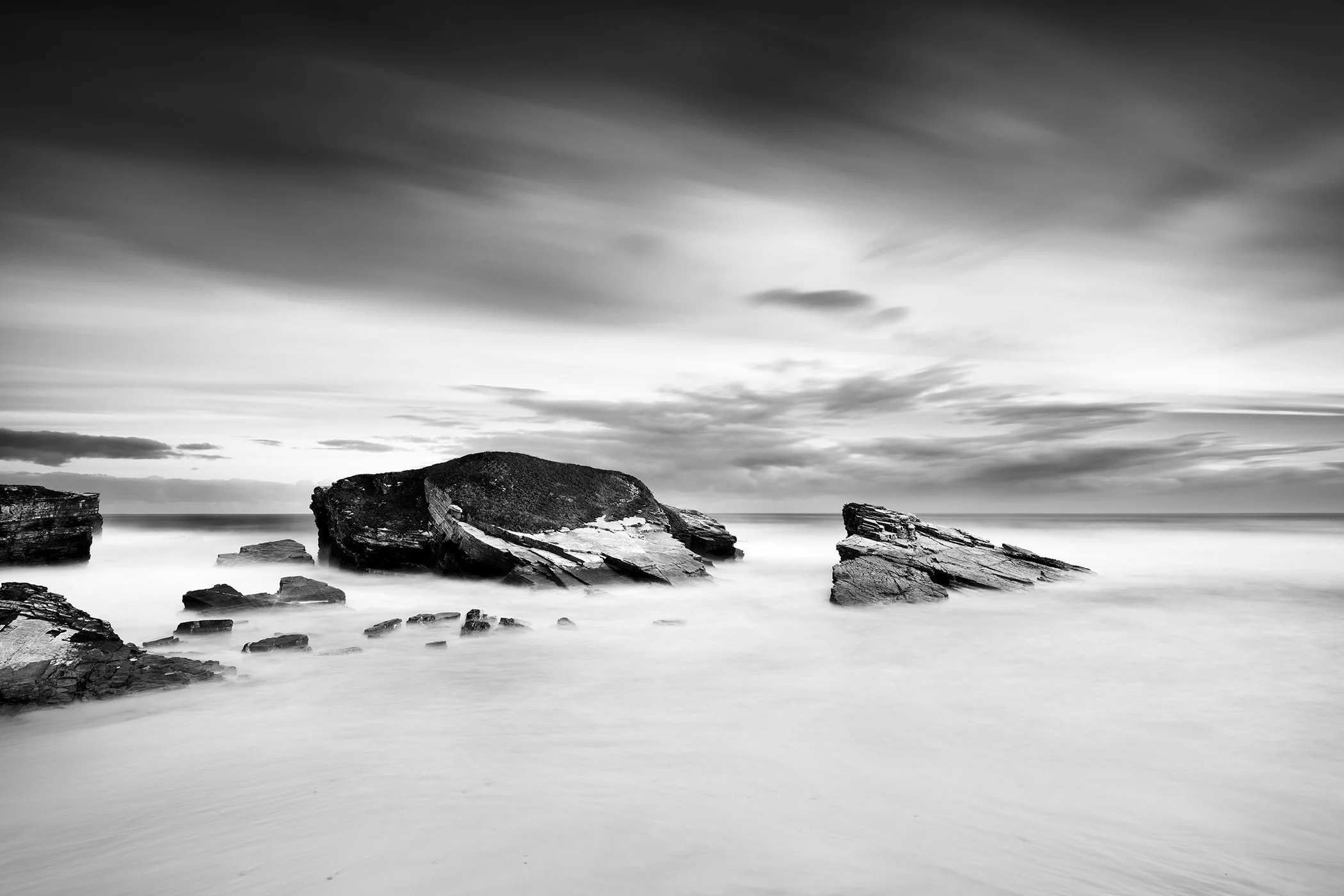 Black-and-white long-exposure seascape of a rocky shoreline with large boulders in calm water under a cloudy sky.