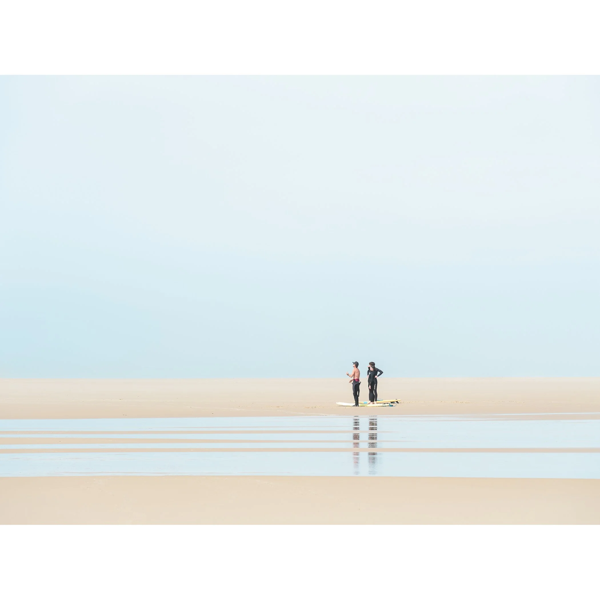 Minimalist colour photograph of three surfers on a quiet beach in France with shallow tidal water and soft reflections. Photo by Gerald Berghammer.