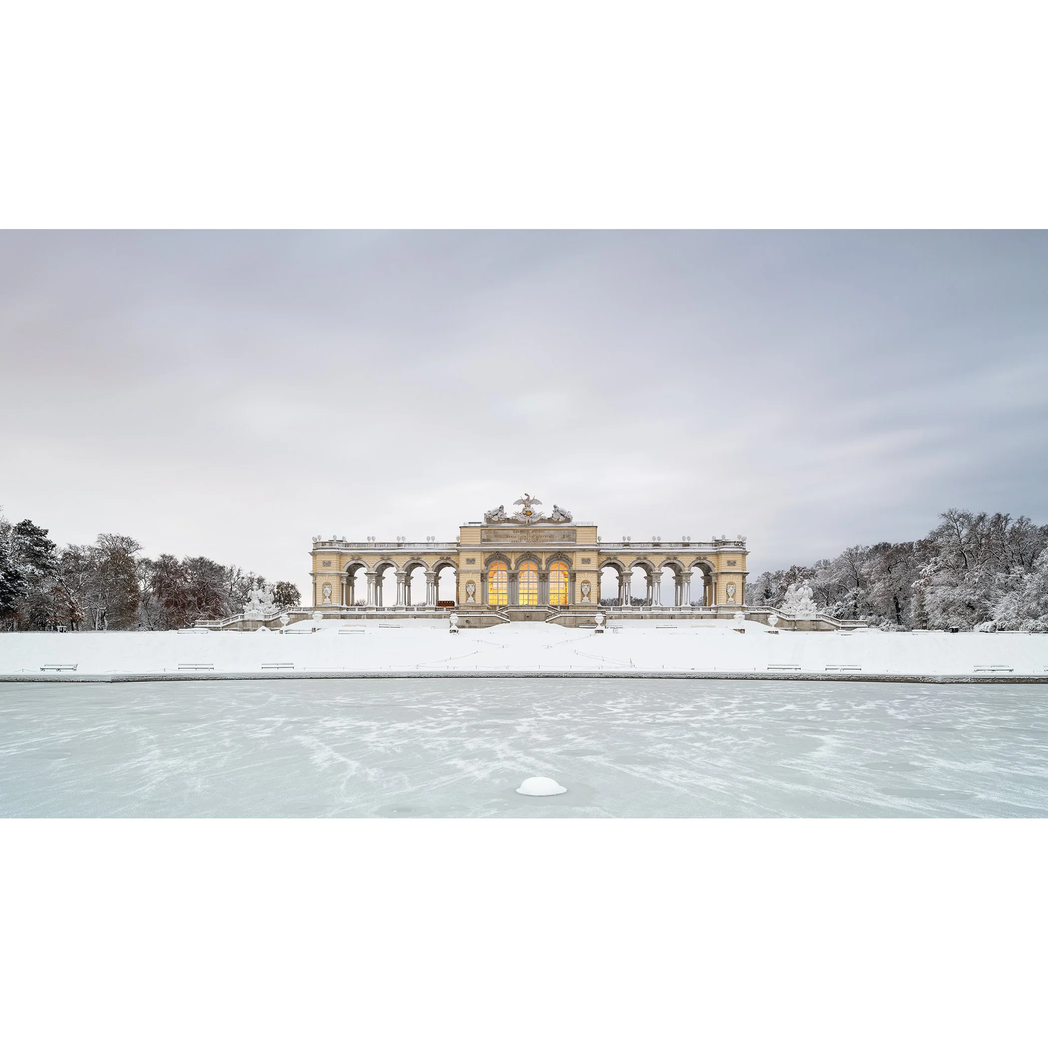 Gerald Berghammer - Color Photography. A historic building on a snow-covered landscape, surrounded by leafless trees, with a frozen body of water.