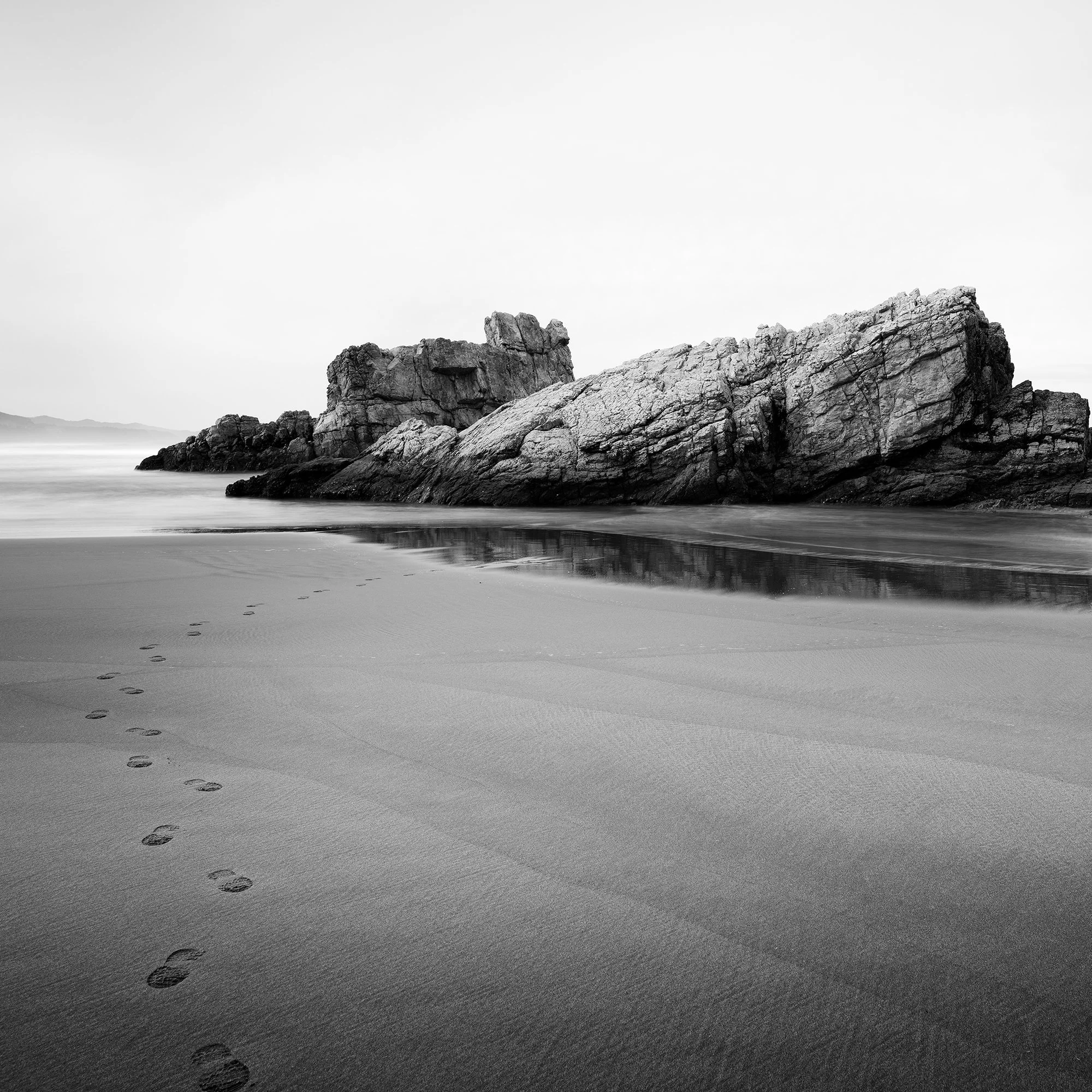 © 2023 Gerald Berghammer. Black-and-white beach scene with wet sand, faint footprints, and a tall rock formation near the shore reflected in shallow water. Print detail 1