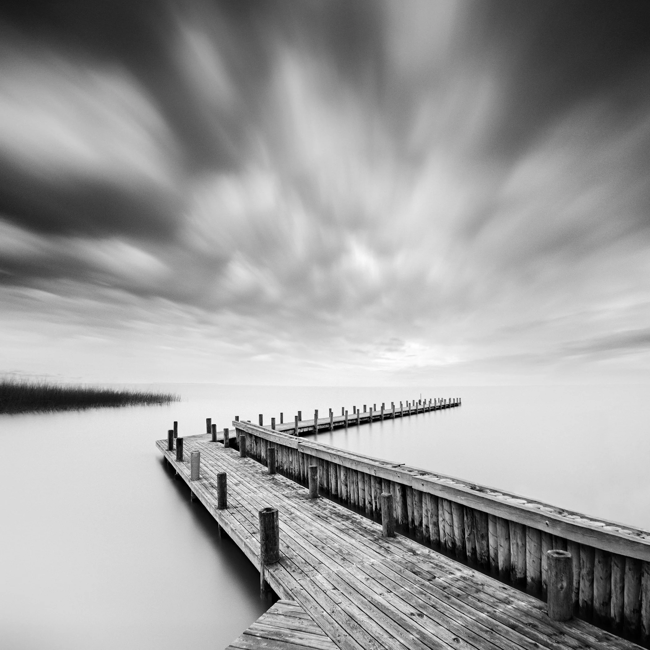 Analogue black-and-white long-exposure photograph of a wooden pier stretching across calm water beneath a cloudy sky.