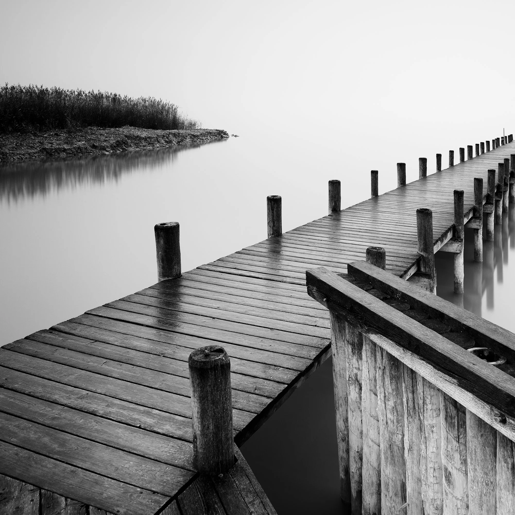 Gerald Berghammer - Black and white photography. Wooden pier extending out over calm water, with a foggy background and some reeds on the shoreline. Print detail 2
