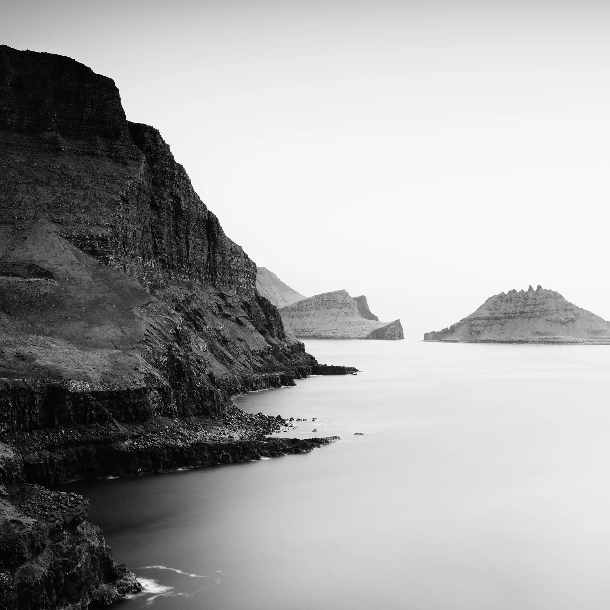 Minimalist monochrome coastal scene with dark cliffs, still water and remote island formations