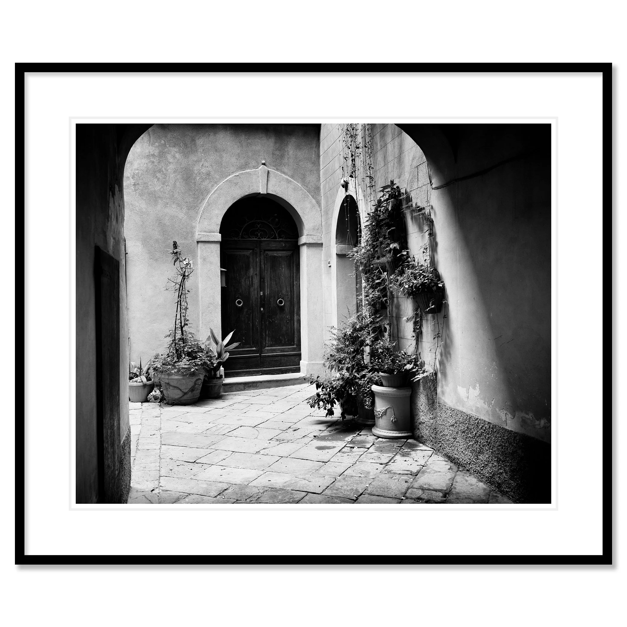 Gerald Berghammer - Black and white cityscape photography. A courtyard with a wooden door, arched doorways, and potted plants on a cobblestone floor. Classic framed black