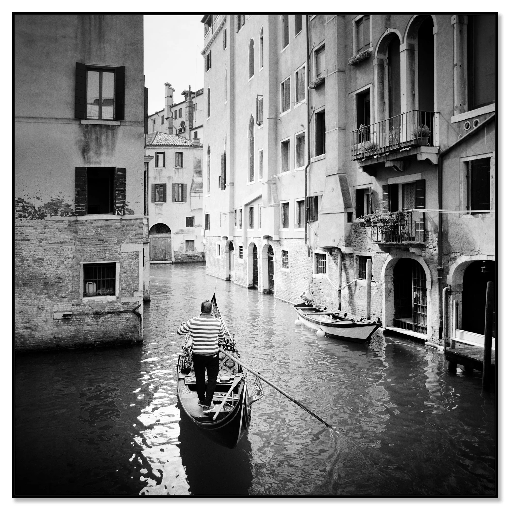 Gondolier rowing a gondola through a narrow canal in Venice, Italy, between tall historic buildings – framed ArtBox black