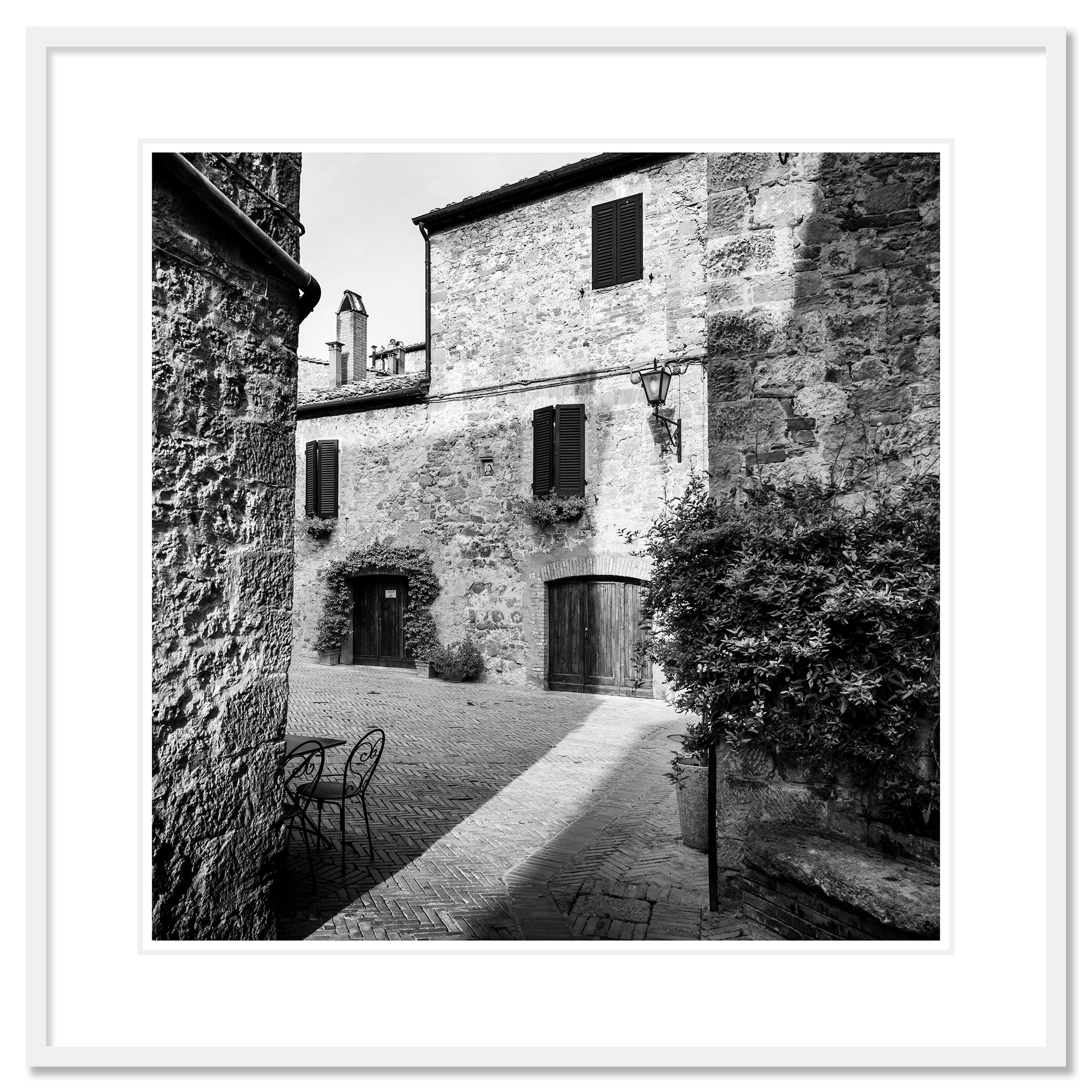 Gerald Berghammer - Black and white cityscape photography. Old stone building with wooden shutters, potted plants and outdoor seating in a narrow courtyard. Classic framed white