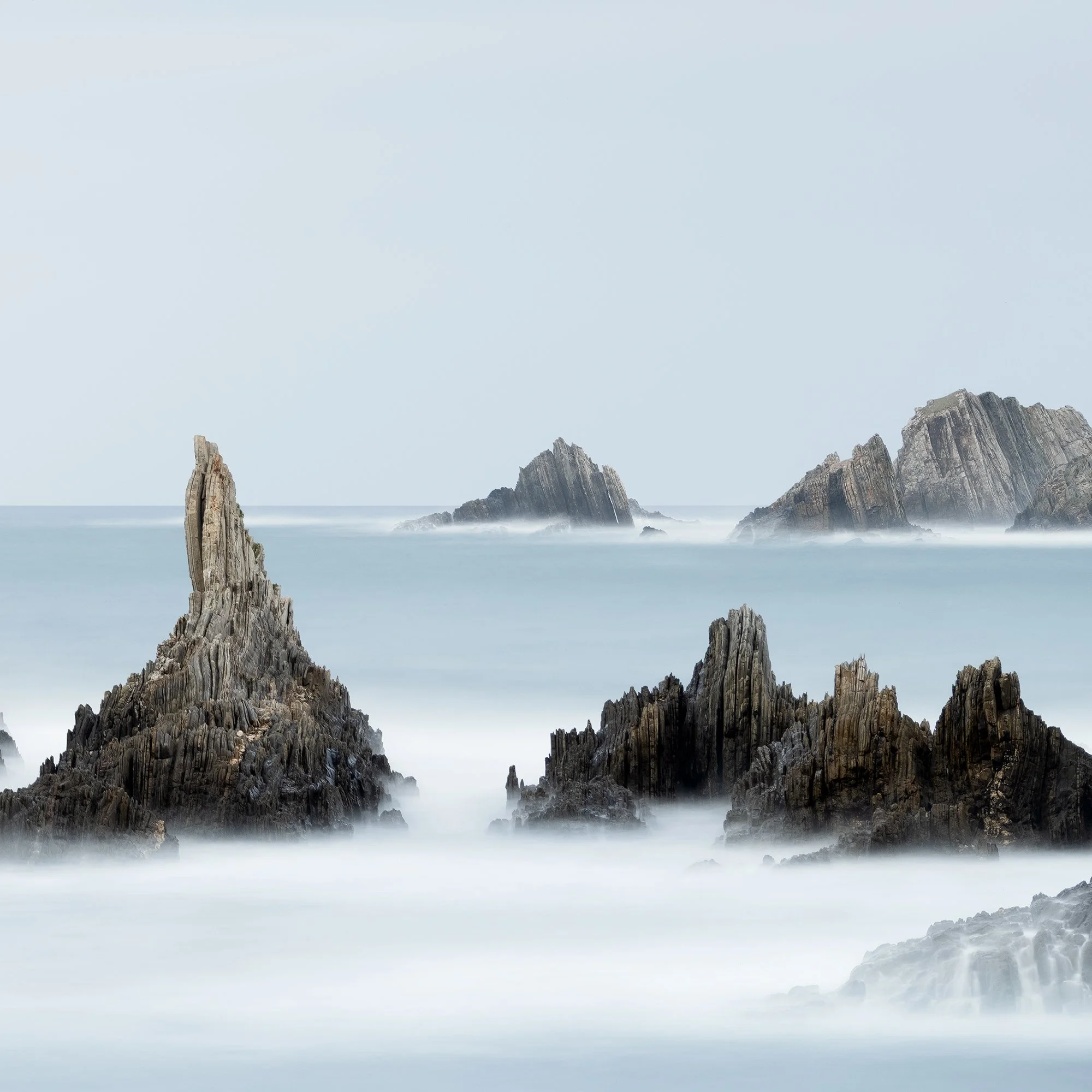 © 2023 Gerald Berghammer - Color seascape. Long exposure photograph of jagged sea stacks rising from the ocean with misty water and a light sky in the background. Print detail 2