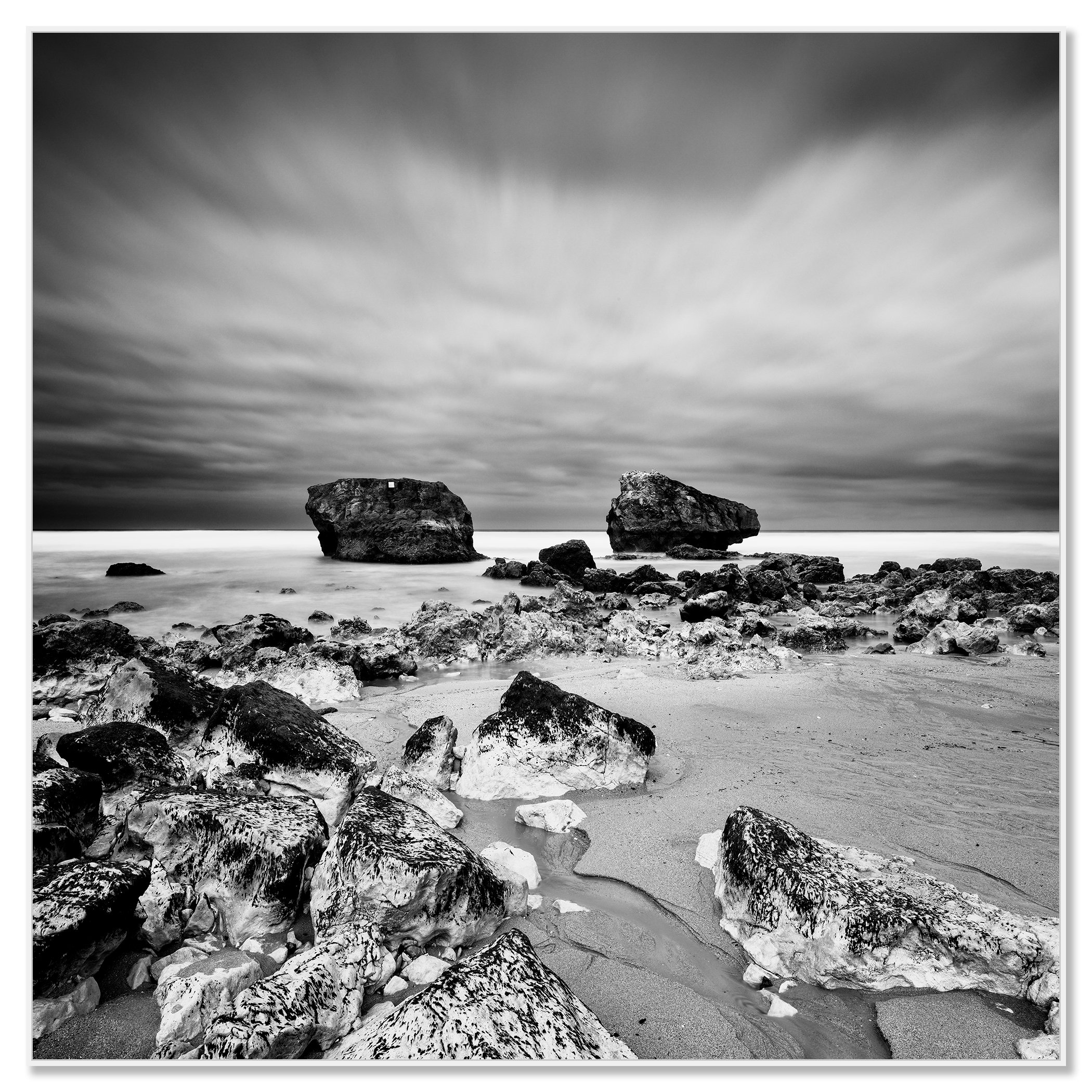 Black-and-white rocky beach with boulders offshore under dramatic, streaked clouds – framed ArtBox white