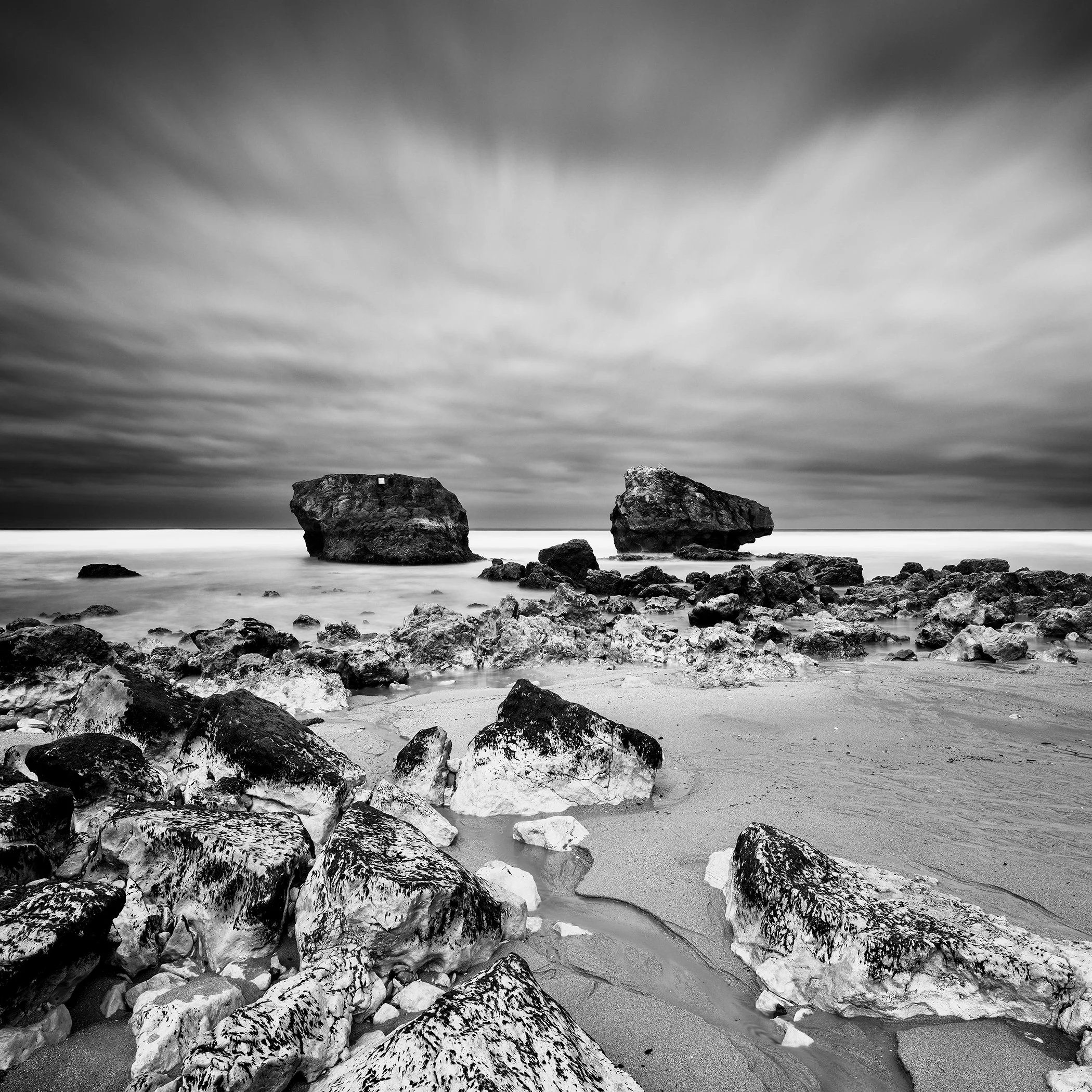 Black-and-white coastal landscape featuring a rugged shoreline, sea rocks, and textured clouds.