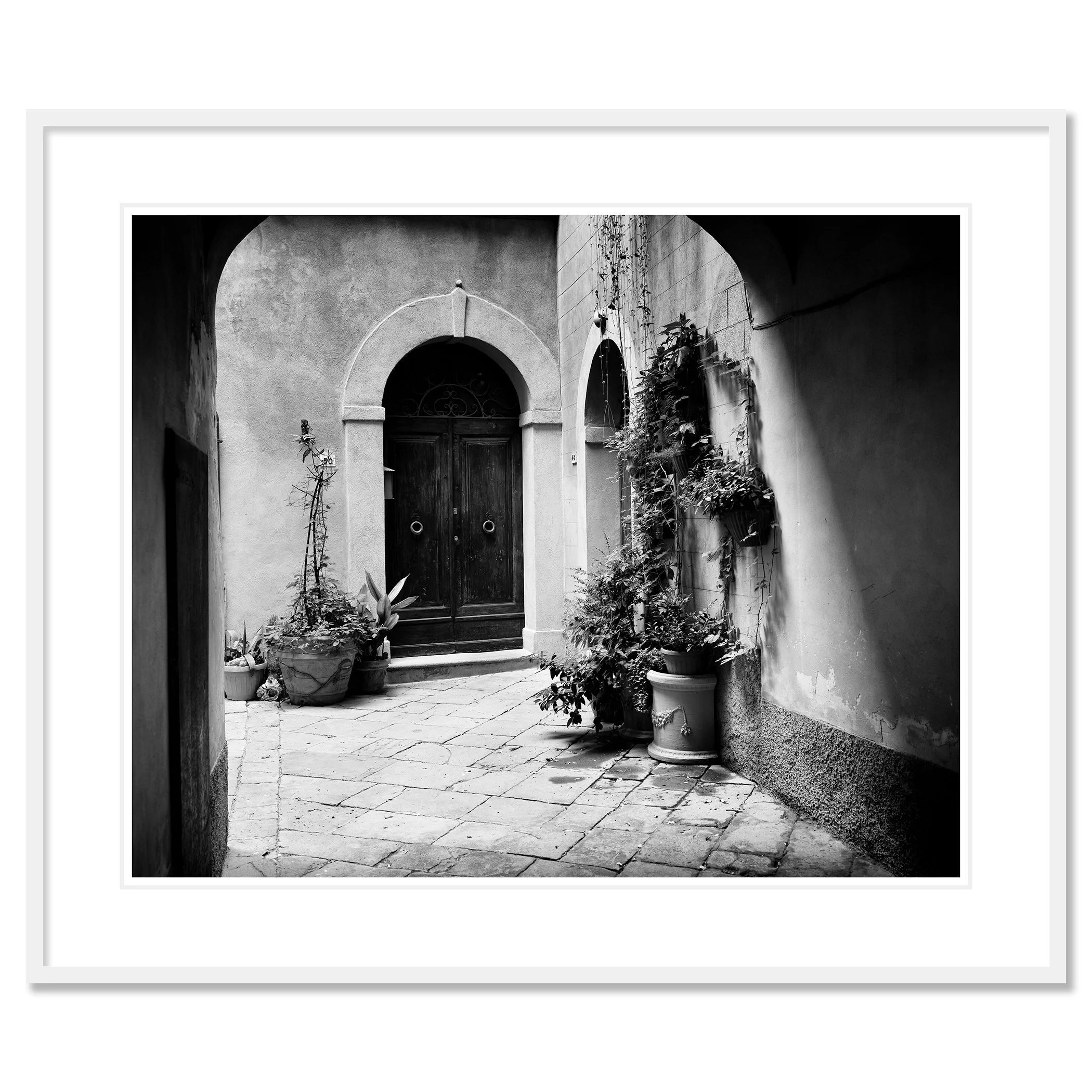 Gerald Berghammer - Black and white cityscape photography. A courtyard with a wooden door, arched doorways, and potted plants on a cobblestone floor. Classic framed white