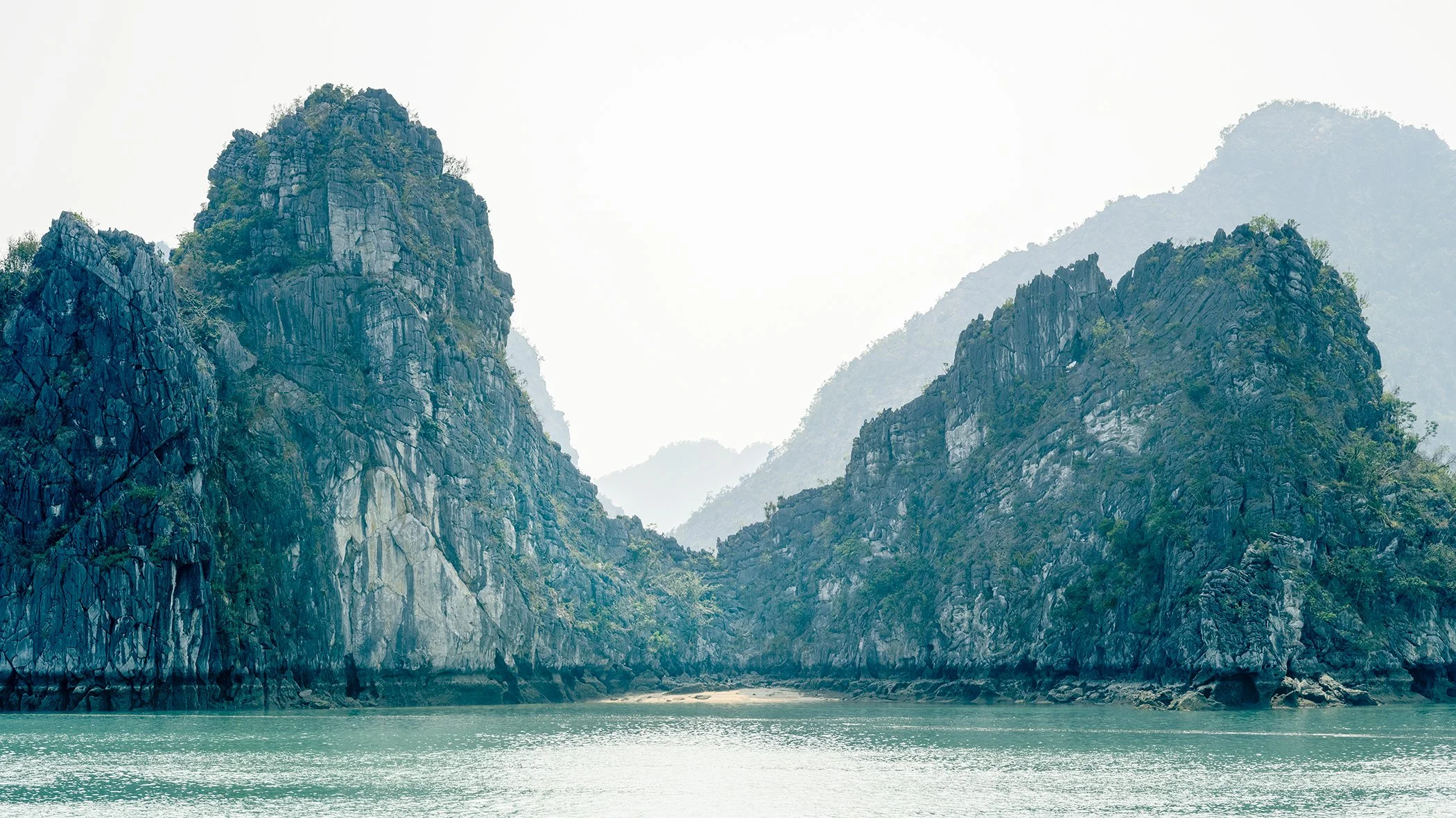 Limestone karst cliffs surrounding a secluded beach above calm turquoise water in Ha Long Bay, Vietnam, photographed by Gerald Berghammer.