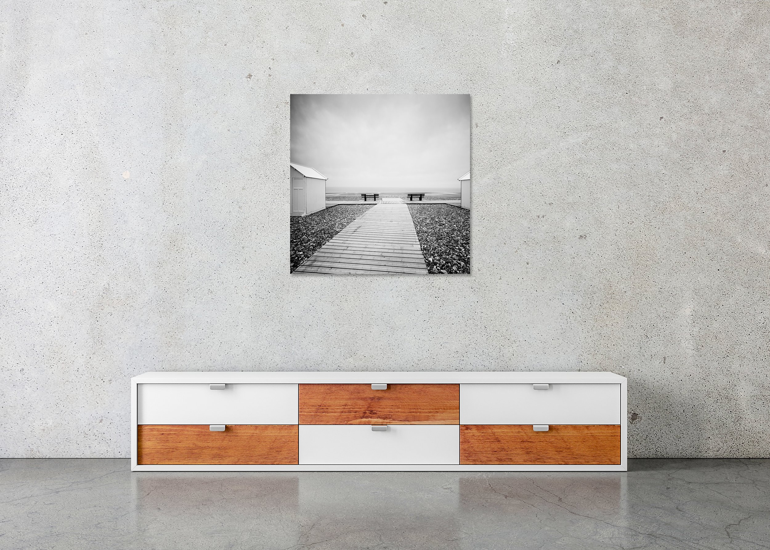 Black and white photo print of a wooden path on a shingle beach between white beach huts, mounted on Dibond with a frameless finish.