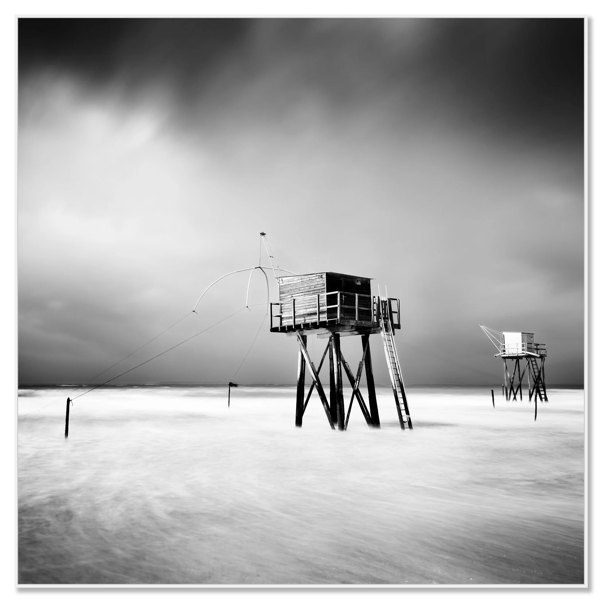 Black and white long-exposure photo of wooden fishing huts on stilts, reached by ladders, over calm sea water under a dramatic cloudy sky – framed ArtBox white