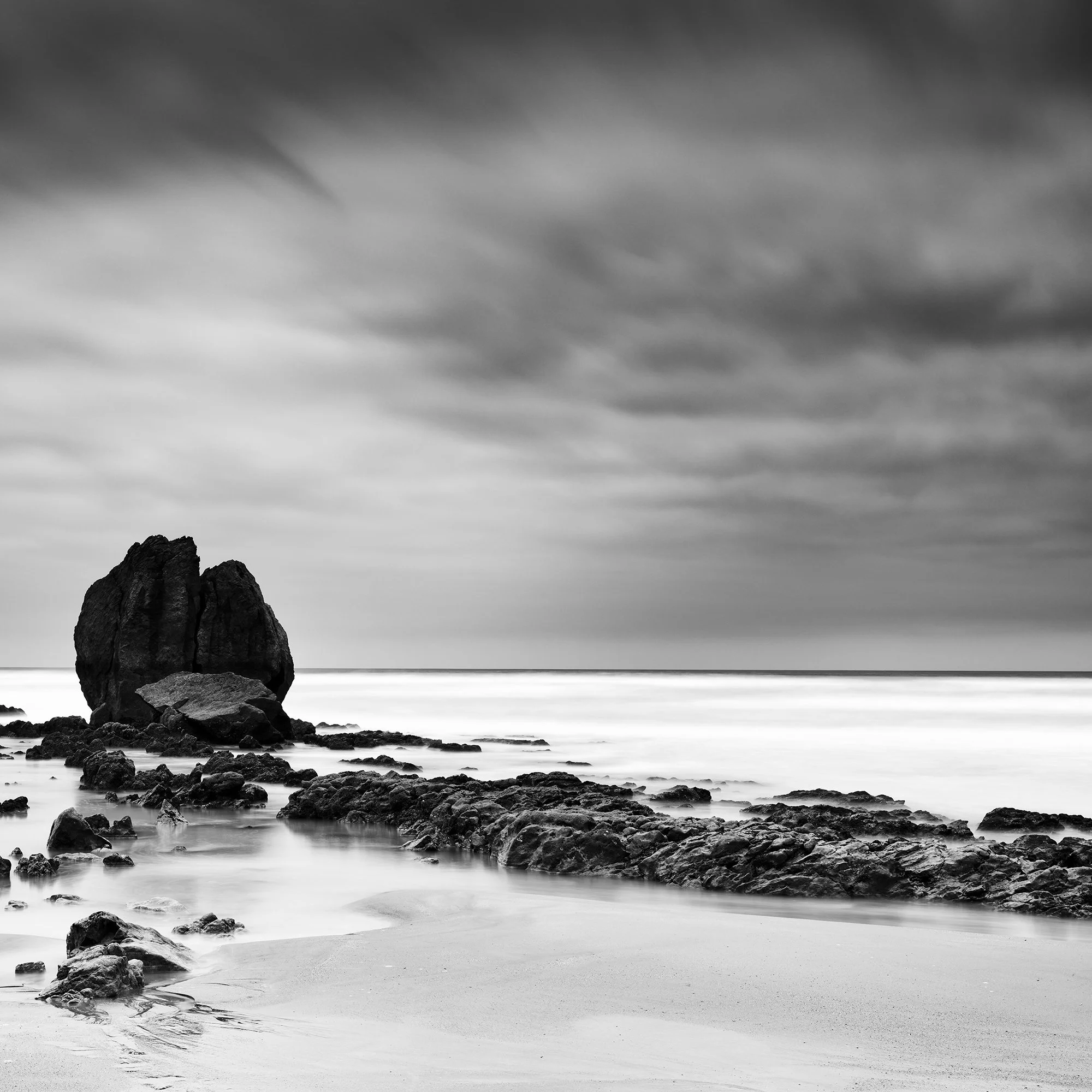 Rocks on the Shore Study 1 by Gerald Berghammer, black-and-white coastal photograph highlighting rock formations, wet sand and an expansive cloudy sky.