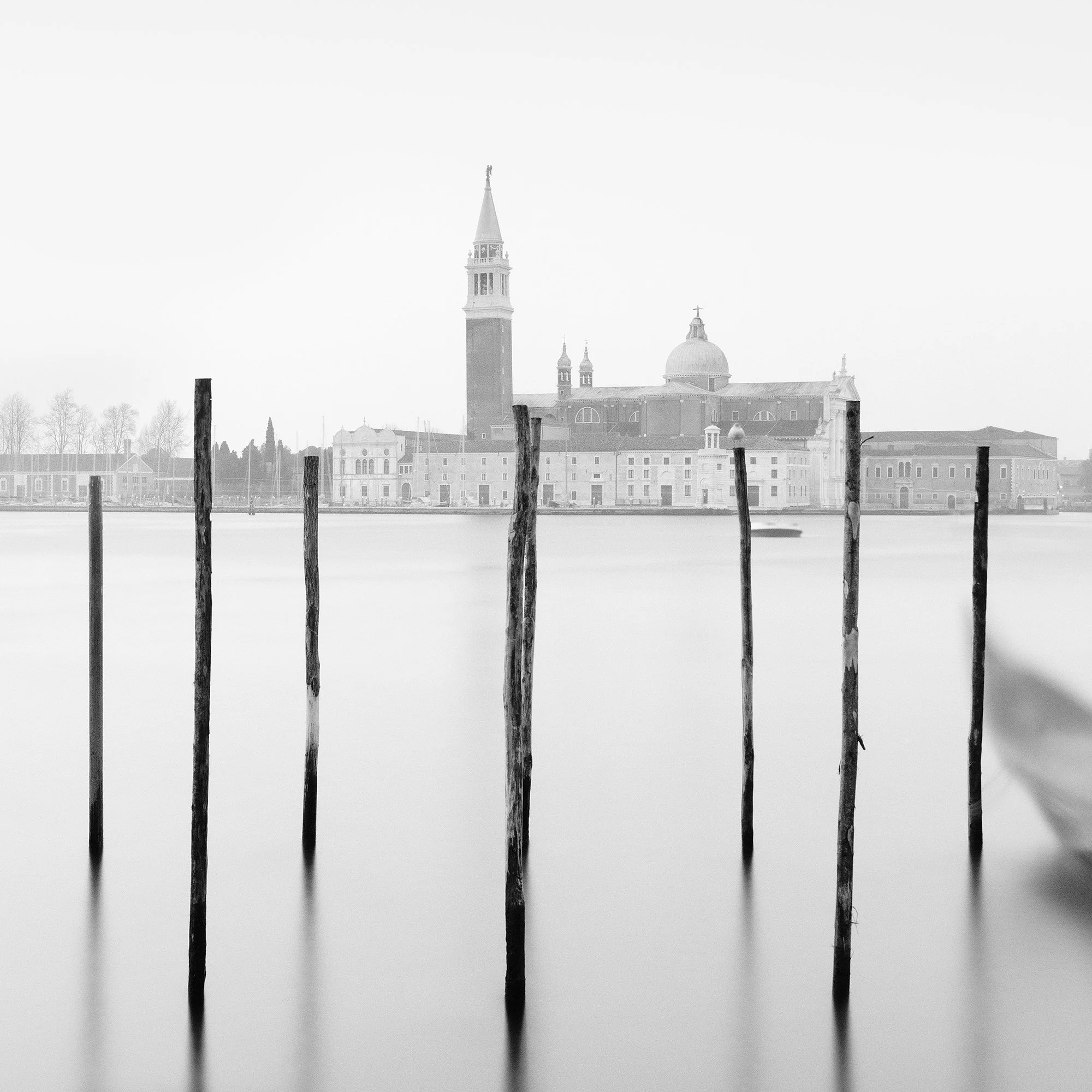 Gerald Berghammer - Black and white cityscape photography. Gondolas moored along wooden posts on a canal with view of a city skyline and a tall bell tower. Print detail 3