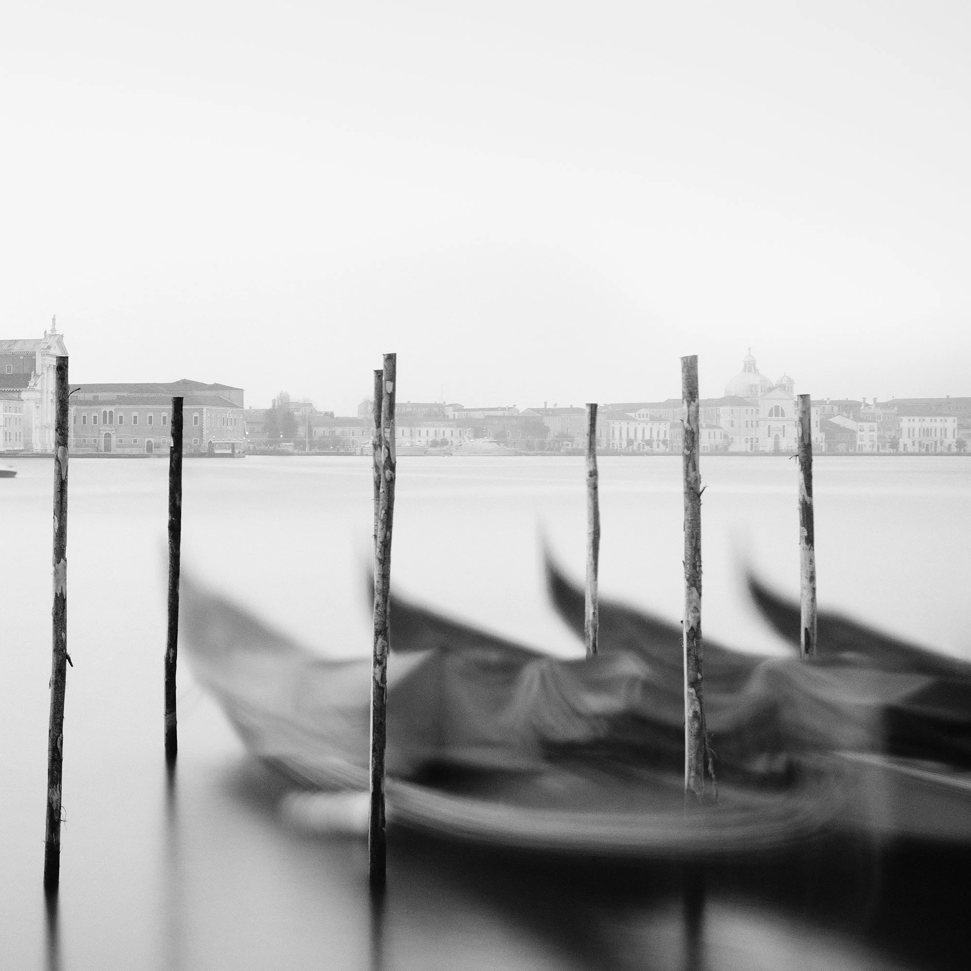 Gerald Berghammer - Black and white cityscape photography. Gondolas moored along wooden posts on a canal with view of a city skyline and a tall bell tower. Print detail 2