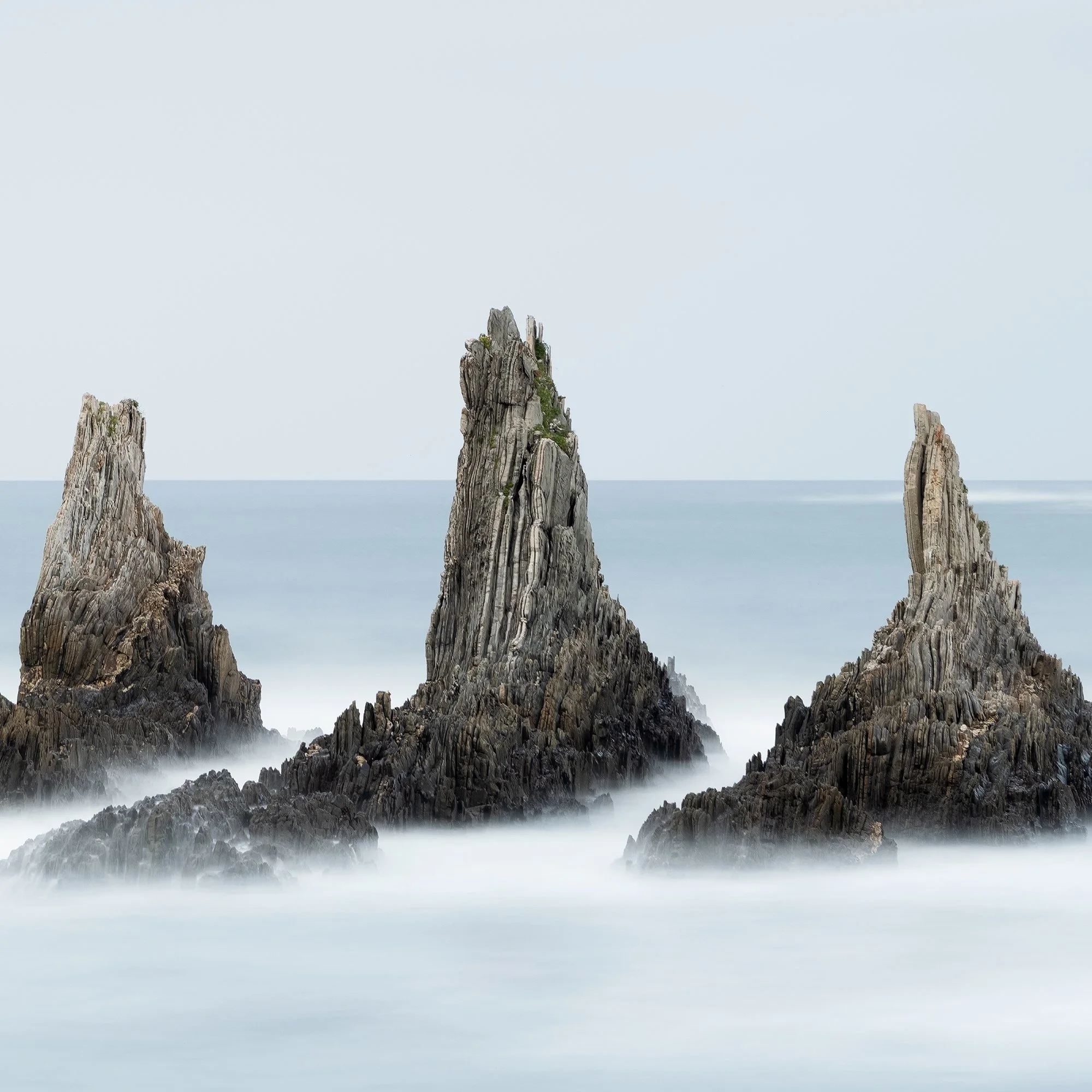 © 2023 Gerald Berghammer - Color seascape. Long exposure photograph of jagged sea stacks rising from the ocean with misty water and a light sky in the background. Print detail 3