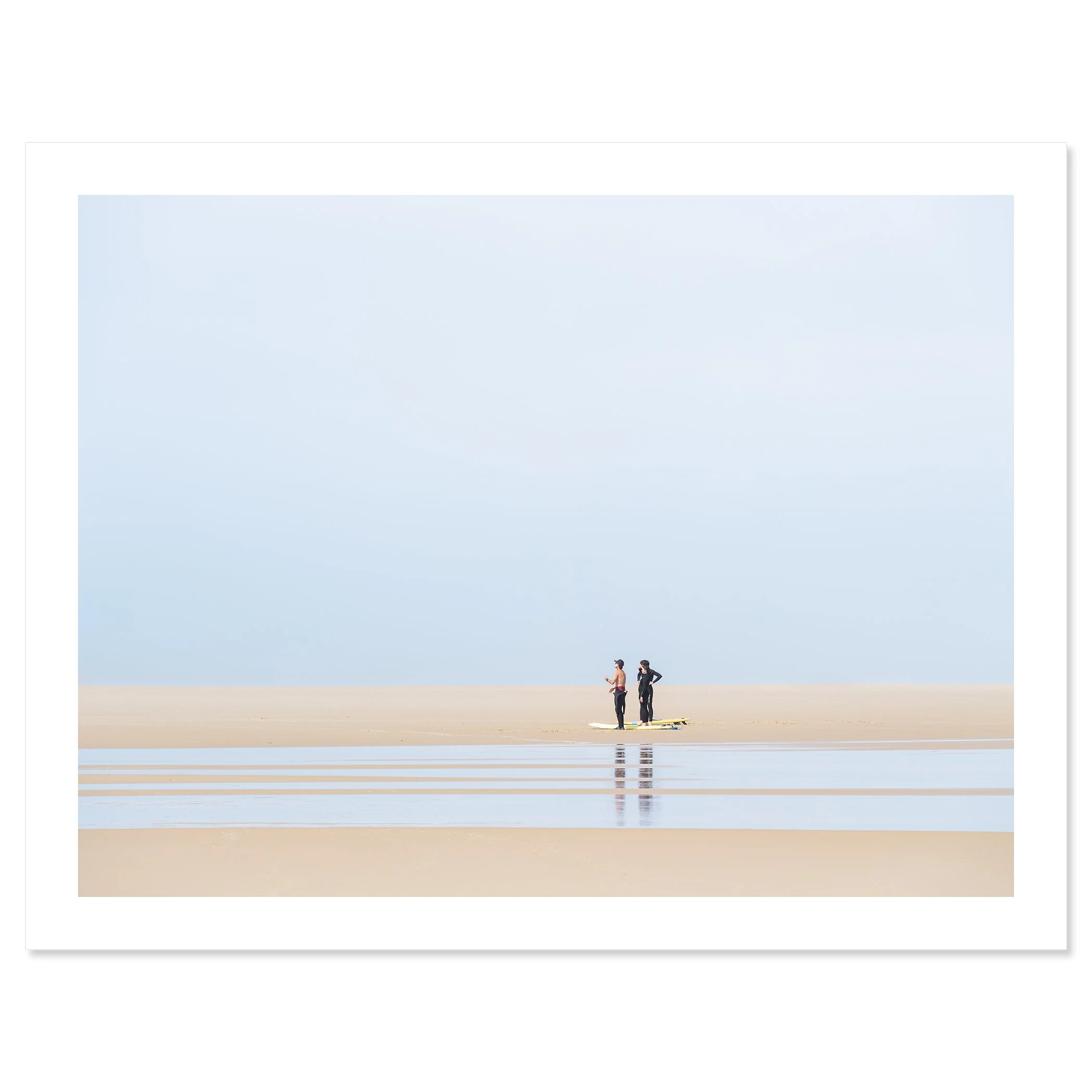 Gerald Berghammer - Color landscape Photography. People stand on a sandy beach near the ocean, wearing wetsuits and surfboards against a pale blue sky. Fine art print only