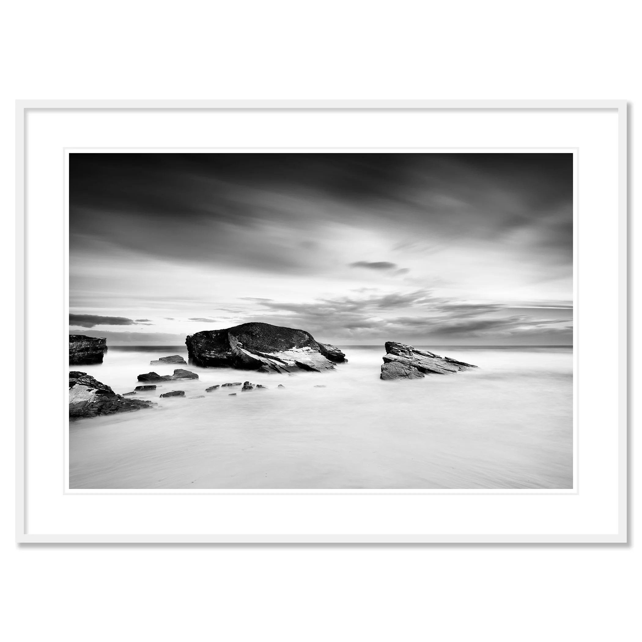 Gerald Berghammer - Black and white long exposure seascape photography. A rocky seashore with large rocks protruding from calm water and a cloudy sky. Classic framed white