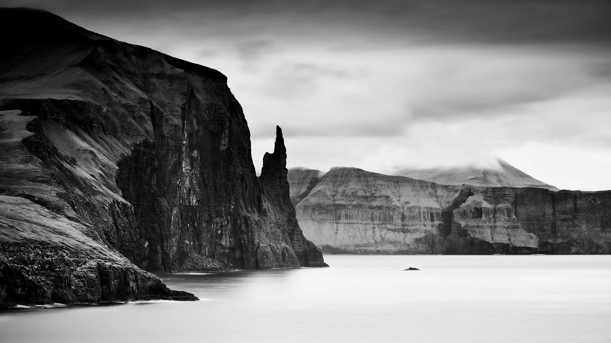 Black and white fine art photograph of dramatic coastal cliffs, a sea stack and calm water under a cloudy sky. Photograph by Gerald Berghammer.