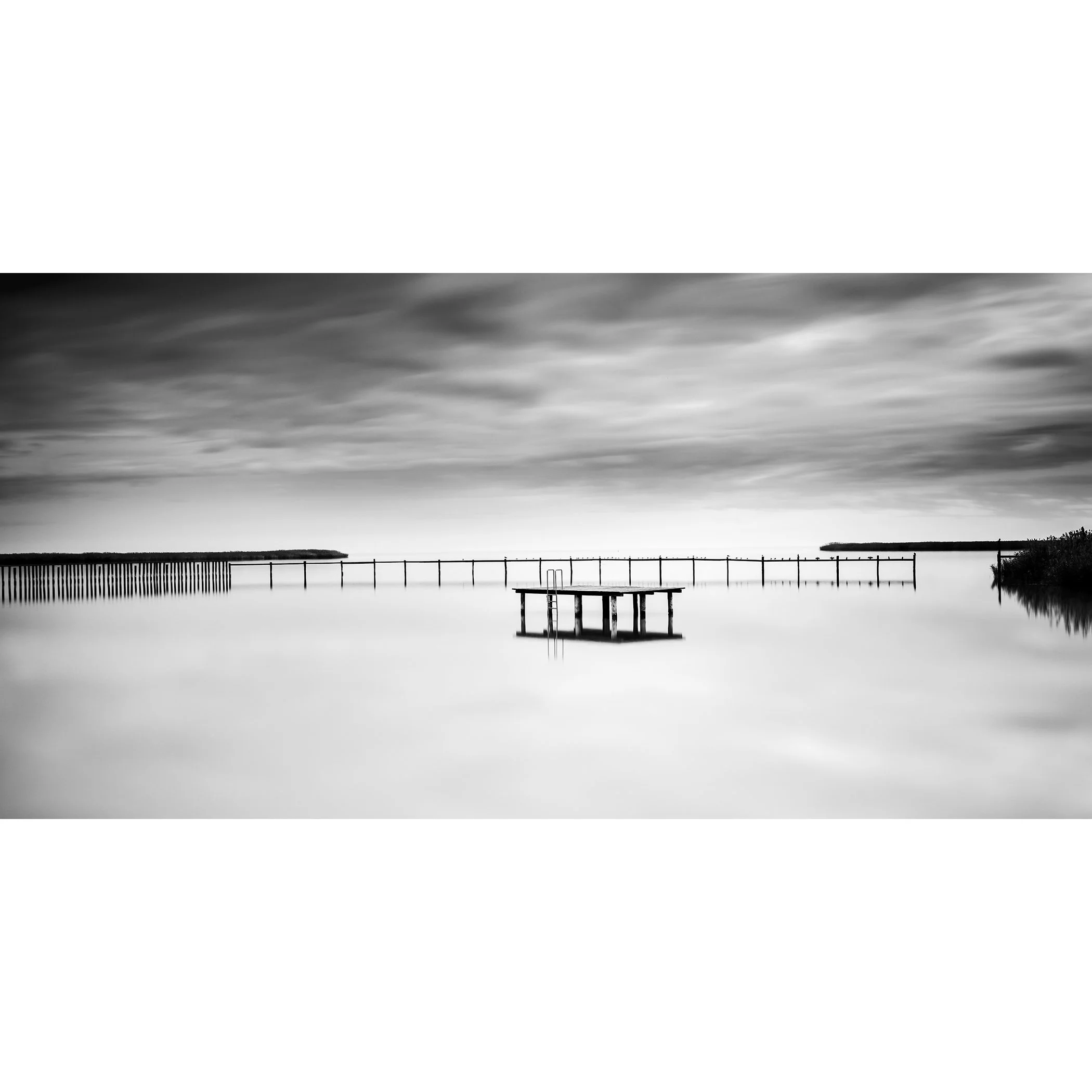© 2023 Gerald Berghammer - Black and white minimalist waterscape. A calm body of water with a floating dock and a pier in the background, under a cloudy sky.