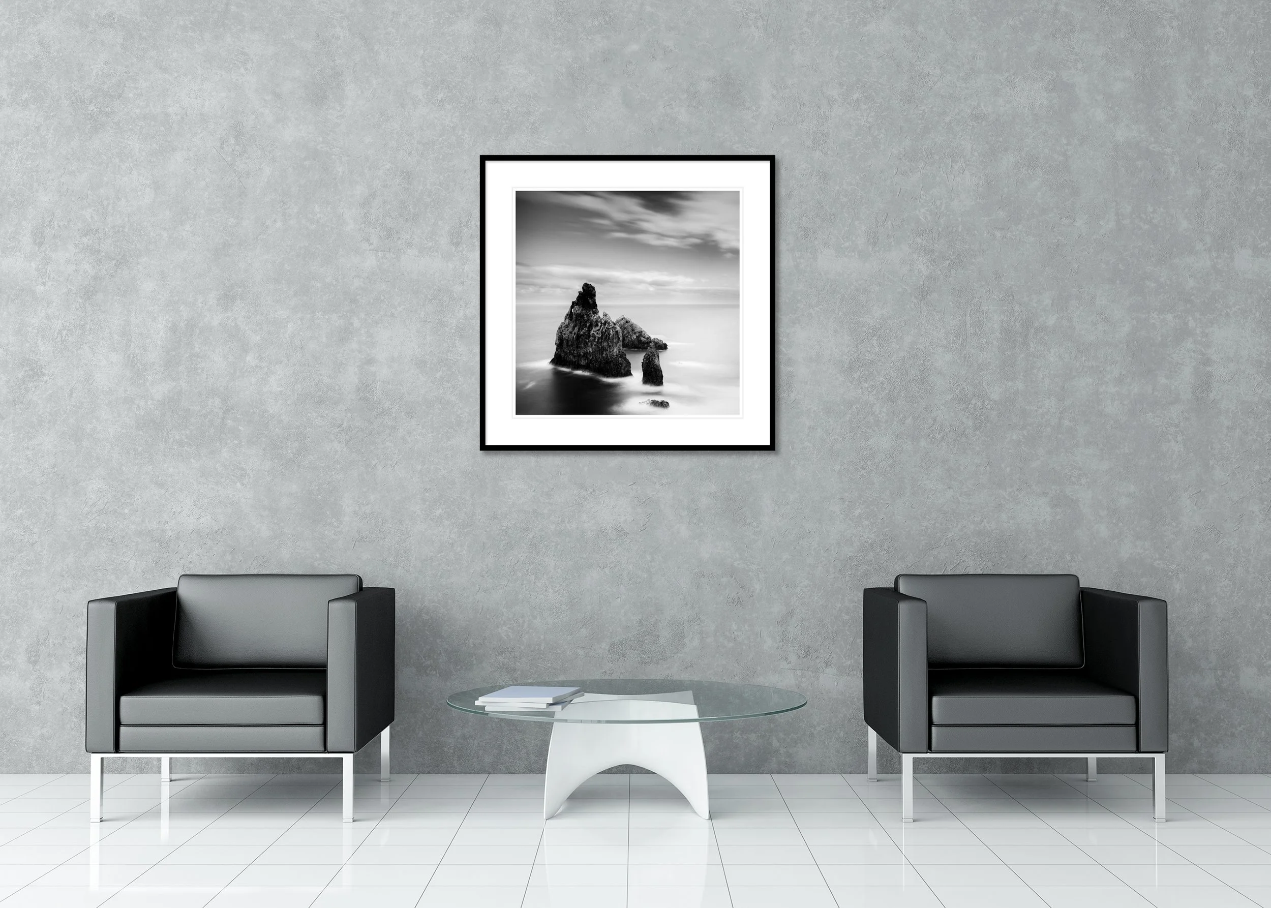 Black-and-white long-exposure photograph of rugged sea stacks emerging from a misty ocean beneath a soft, clouded sky, presented as an archival pigment print in a classic frame with passe-partout.