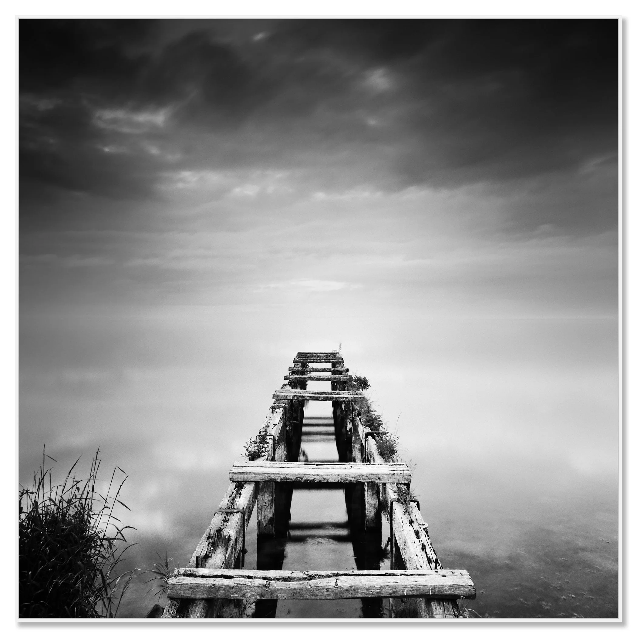 Gerald Berghammer - Black and white waterscape photography. An old abandoned wooden dock extending into a calm body of water under a cloudy sky. Chromaluxe framed white