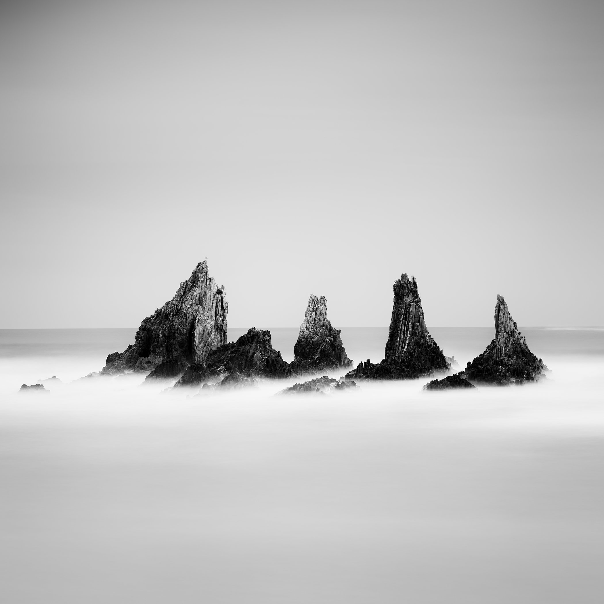 Monochrome seascape showing five rugged sea stacks emerging from the Atlantic, mirrored by a smooth, calm water surface beneath a clear sky.