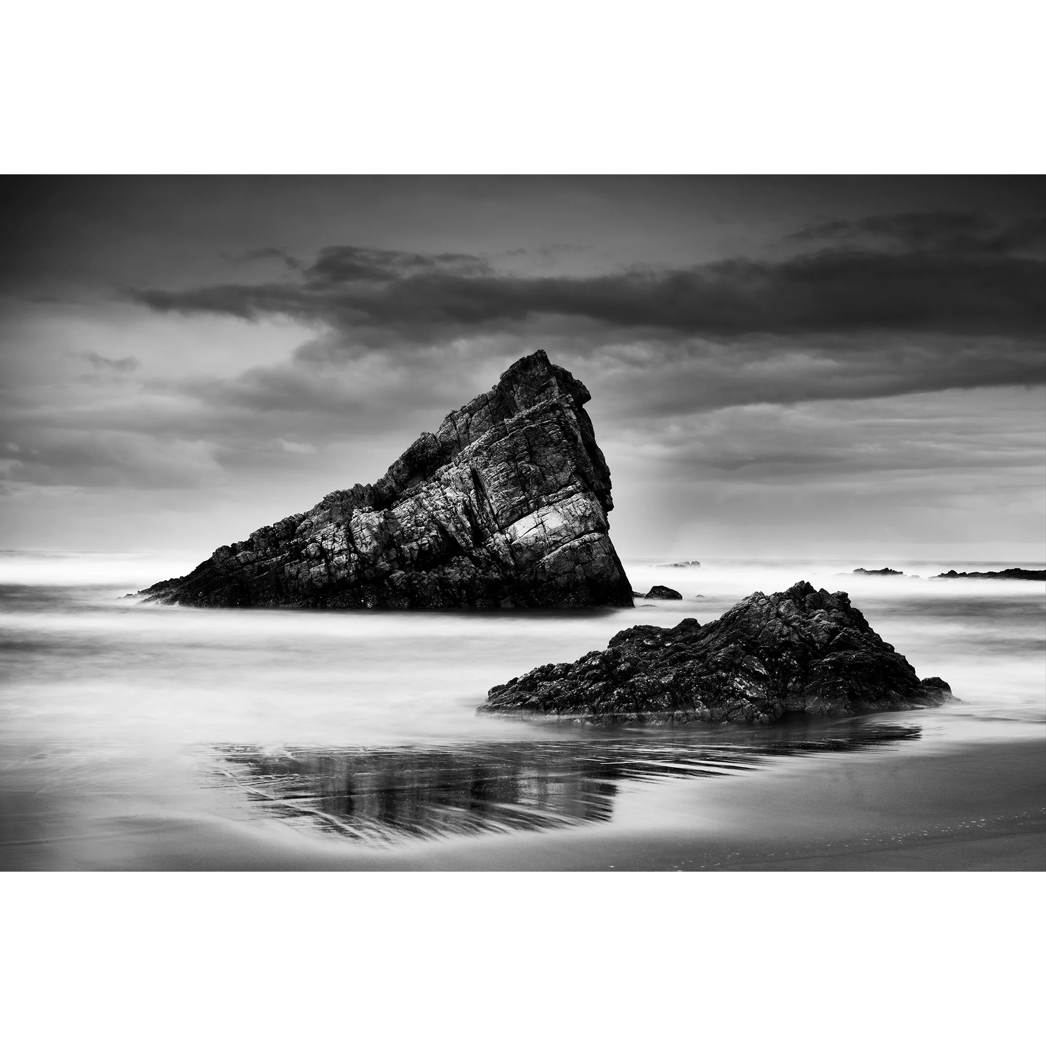 © 2023 Gerald Berghammer. Black-and-white photograph of jagged sea rocks emerging from calm water under a cloudy sky.