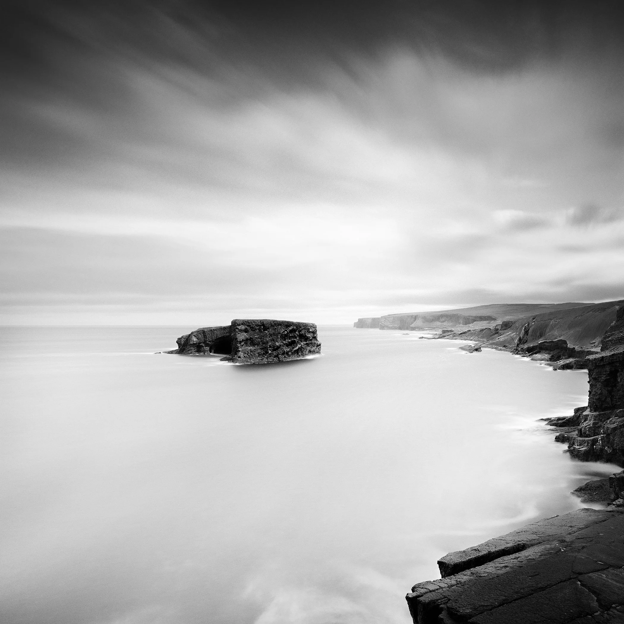 Gerald Berghammer - Black and white long exposure seascape photography. Rocky coastal landscape with cliffs and a large rock formation in the ocean.