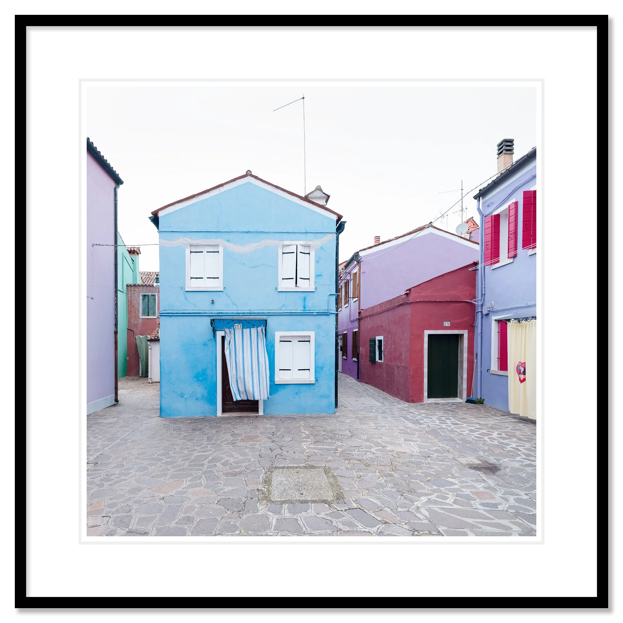 © 2025 Gerald Berghammer photo - Colorful houses in pastel shades on a cobblestone street, with colorful windows and a striped curtain hanging in front of one house. Classic framed black