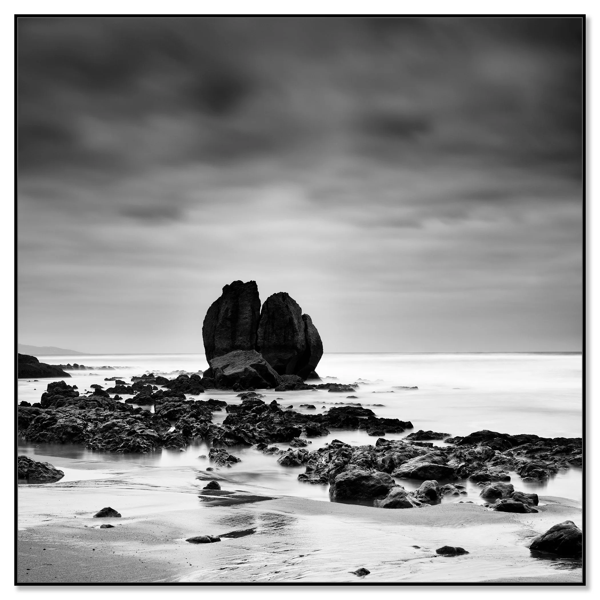 Black and white seascape with a large rock formation on a quiet beach under a cloudy sky – framed ArtBox black