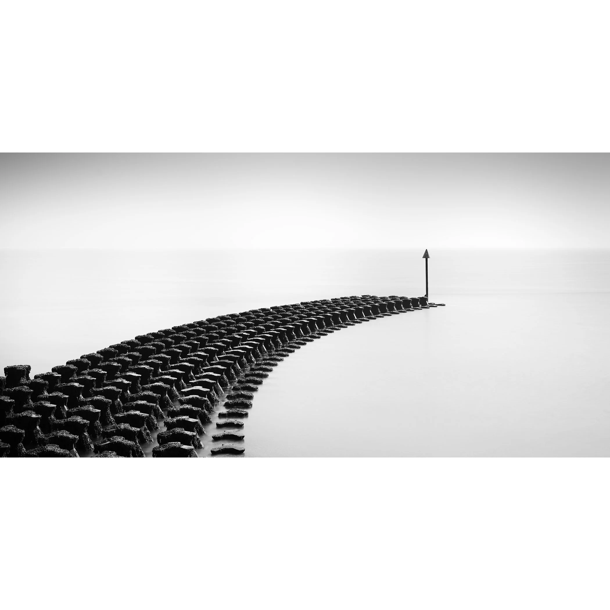 Gerald Berghammer - Black and white minimalist seascape photography. A concrete breakwater with a single arrow marker at the end of the calm sea.