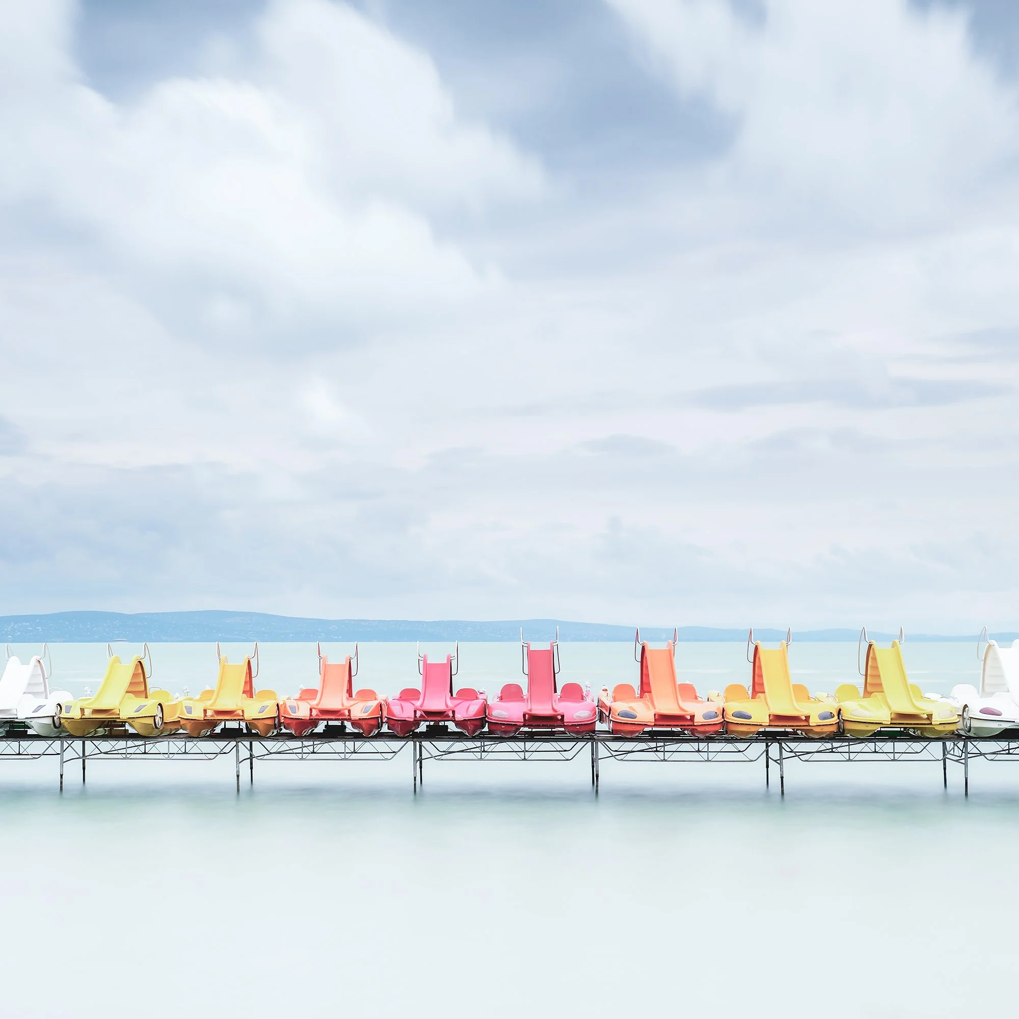 Gerald Berghammer - Color long exposure waterscape photography. Colorful paddle boats lined up on a dock over calm water, under a cloudy sky. Print detail 3