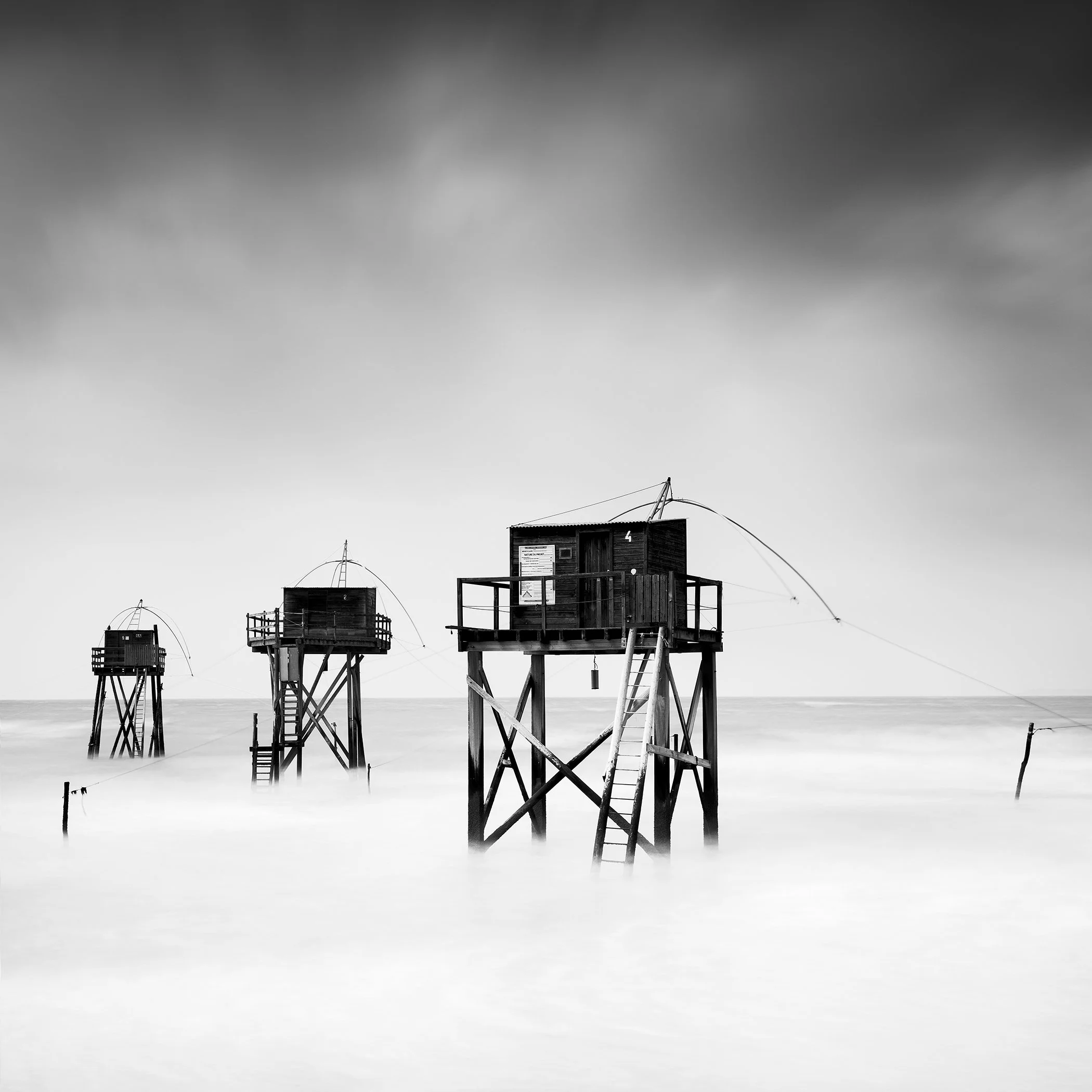 Traditional wooden fishing huts rising above the calm sea in France, photographed in black and white by Gerald Berghammer.