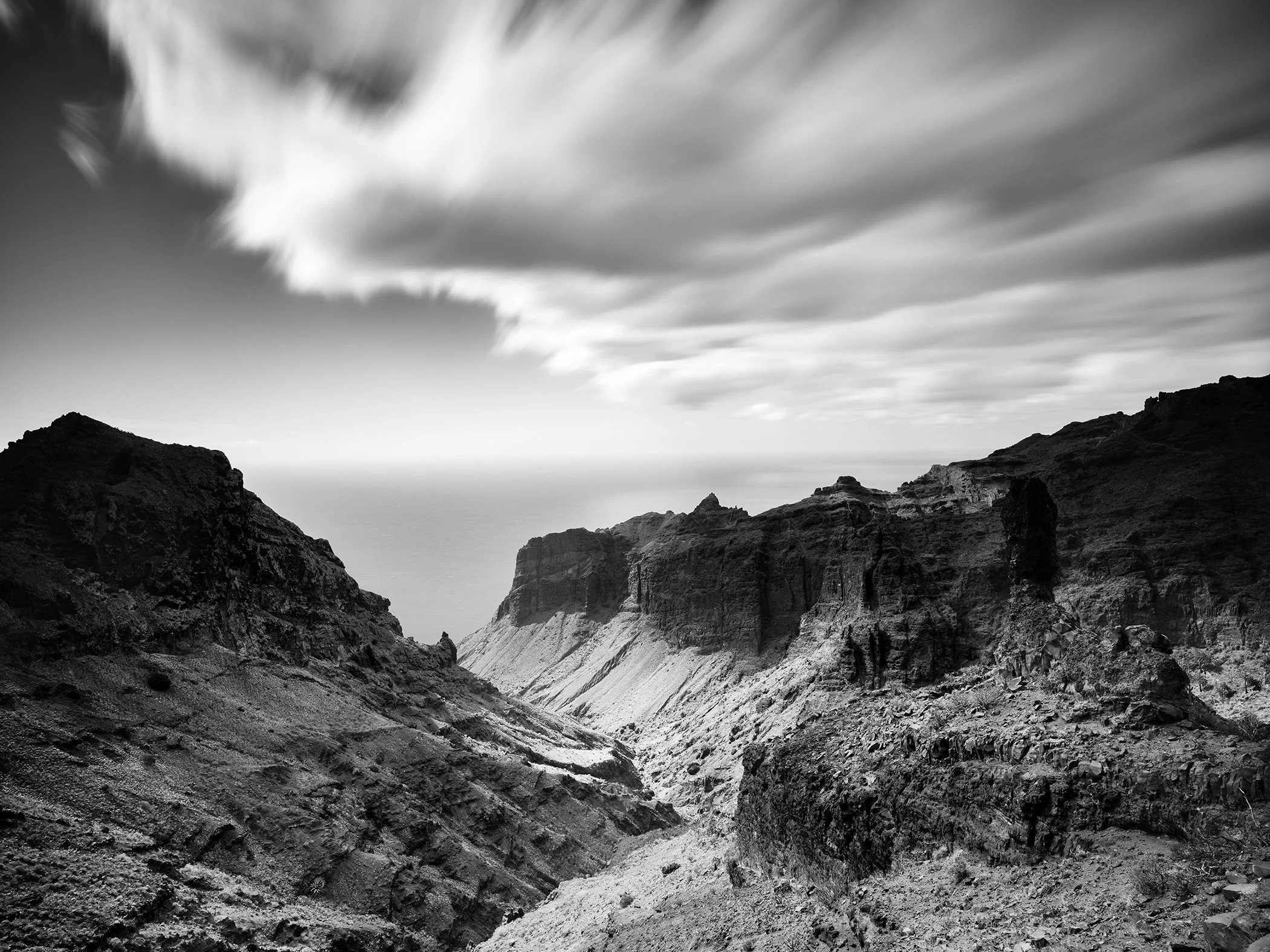 Black-and-white photo of a rugged rocky canyon beneath sweeping clouds, opening towards a distant sea horizon.