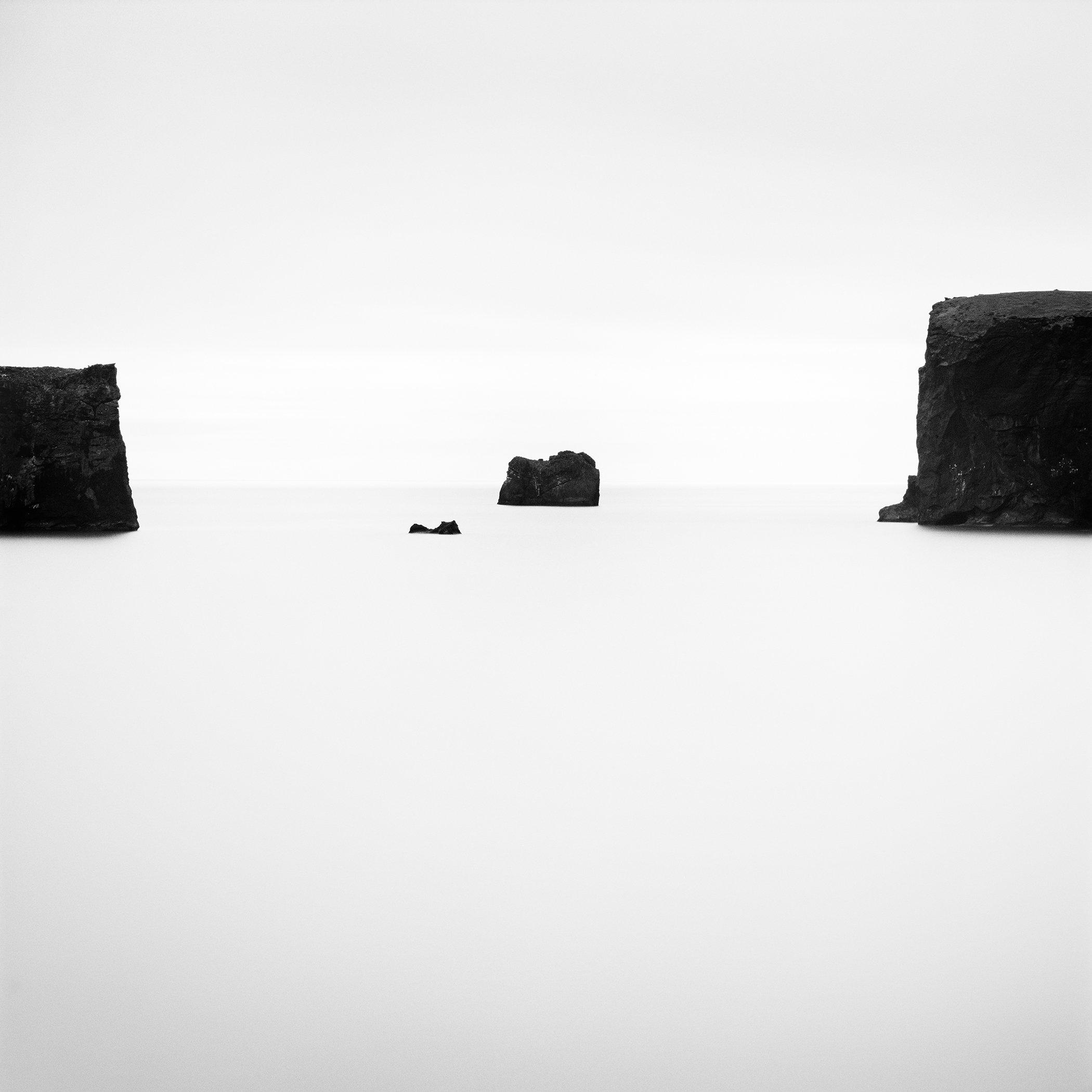 High-contrast black-and-white Icelandic seascape with isolated basalt rocks and cliffs beneath an empty sky. Photo by Gerald Berghammer.