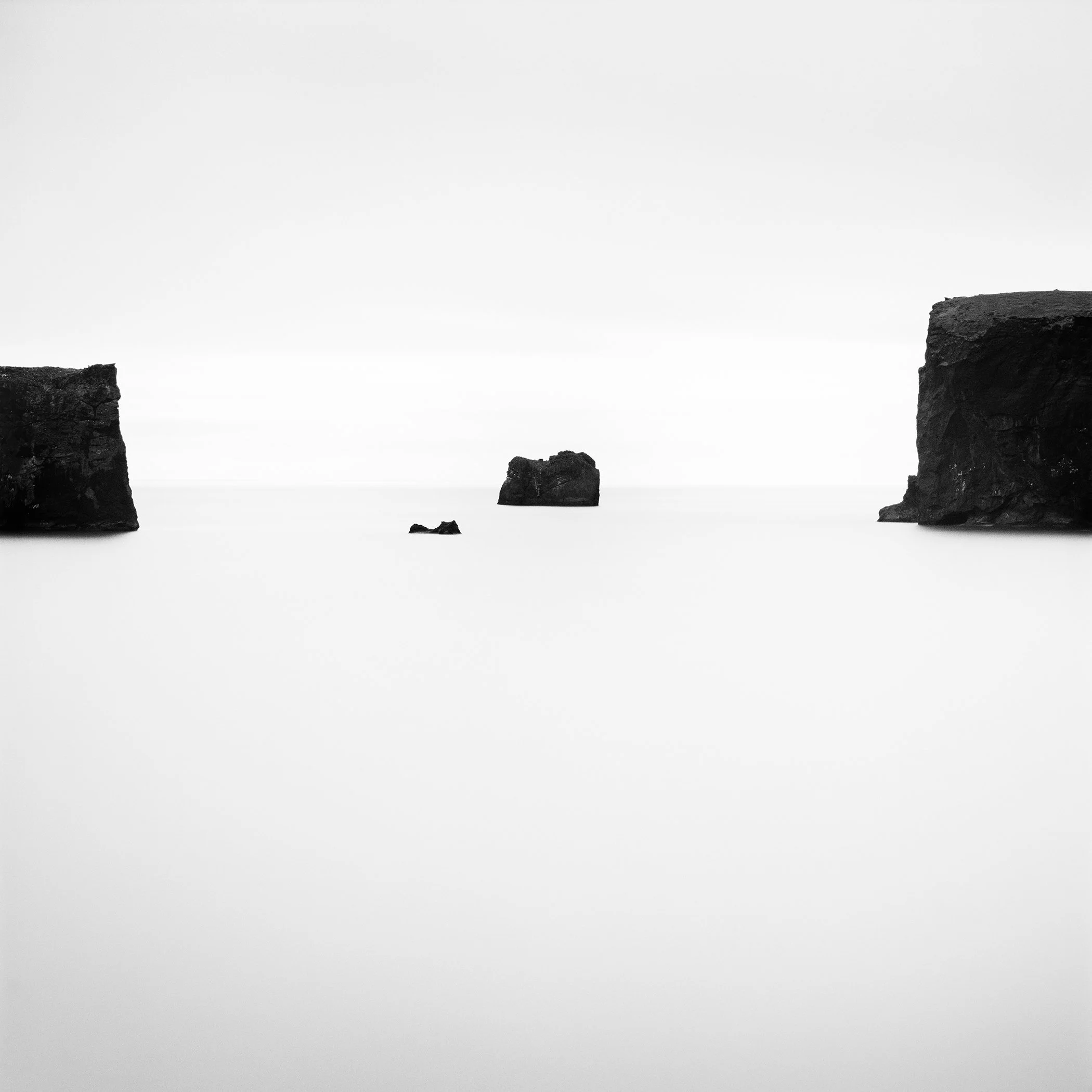 High-contrast black and white minimalist seascape showing calm ocean water with isolated rock formations and cliffs under an empty sky.